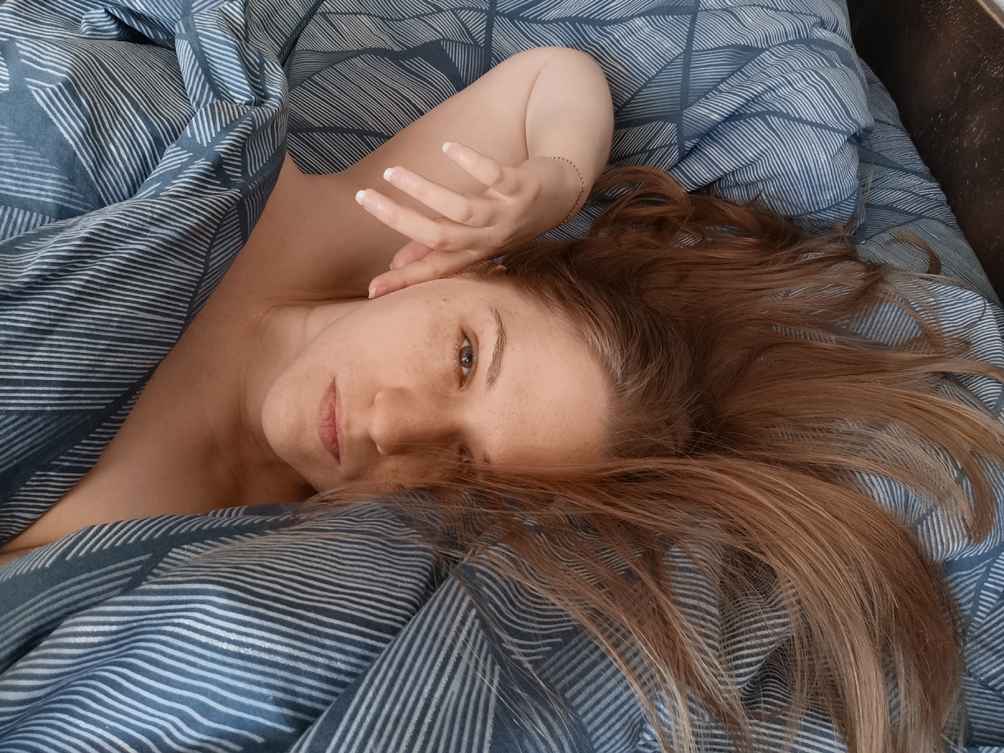 A woman with long, auburn hair lies on her side in bed, covered by blue and white striped bedding. Her left arm is bent and rests near her head, with her hand near her face. Her right arm is hidden beneath the bedding. The woman is looking towards the camera with a neutral expression. The scene is indoors, likely a bedroom. The lighting appears natural, casting soft shadows on her face and body.
