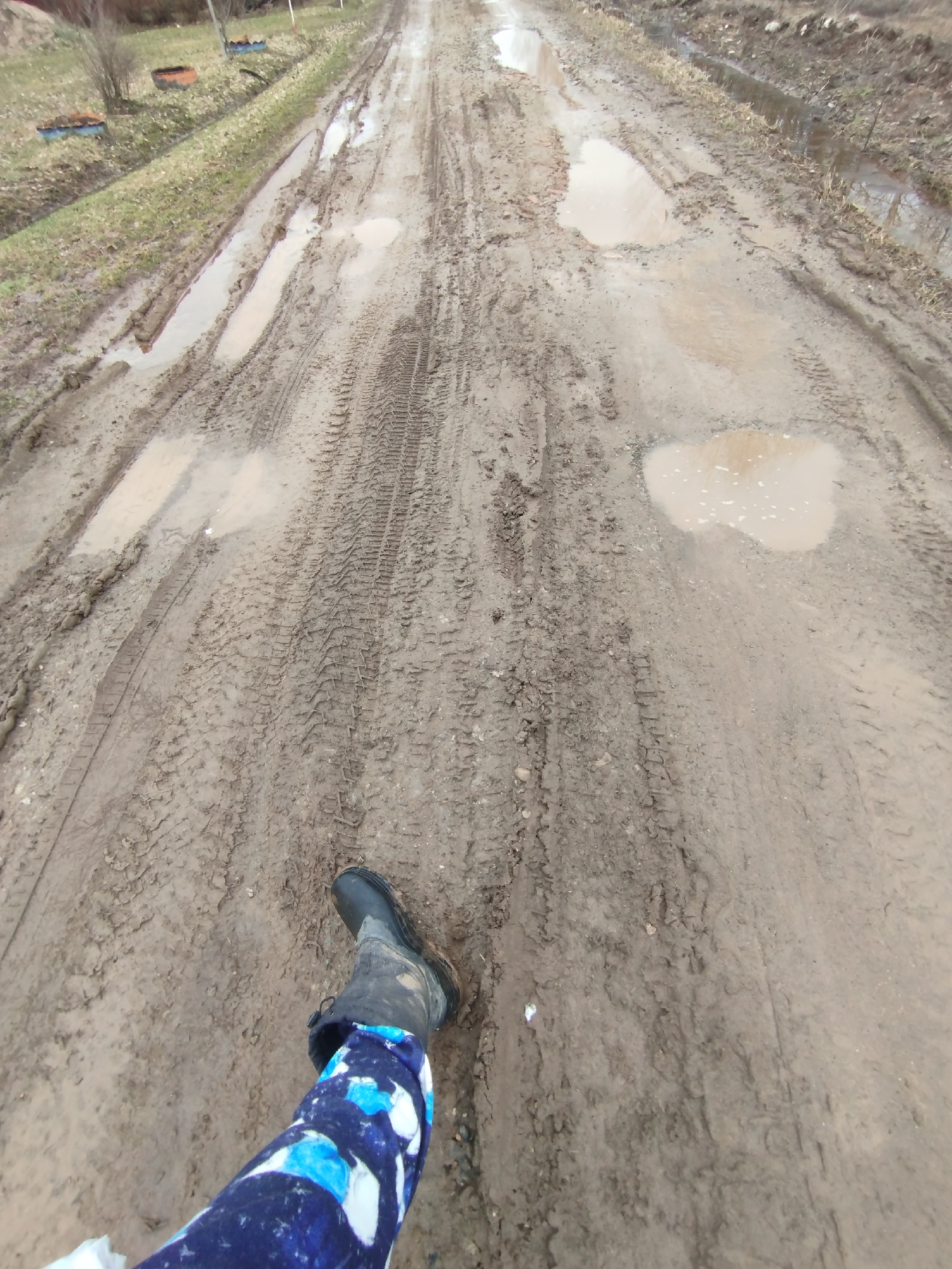 A low-angle shot captures a person's leg and foot navigating a very muddy, unpaved road in Grishanki, Russia. The person is wearing a dark, tall rubber boot on their right foot, with their lower leg covered by pants featuring a blue and white abstract pattern. The foreground and midground are dominated by the dirt road, which is heavily churned with deep tire tracks and numerous large, shallow puddles reflecting the cloudy sky.
Along the left side of the road, a strip of sparse, light green grass separates the track from some bare bushes and small trees in the background. Several overturned buckets or containers, one blue and one reddish-brown, are visible on the grassy verge. The road stretches into the distance, appearing equally muddy and wet. On the right side, the road also features puddles and tire marks, with some indistinct vegetation further back.
The overall scene suggests a rural or semi-rural environment, likely experiencing wet weather conditions such as recent rain or thawing snow, making the path difficult to traverse. The light appears overcast, indicating daytime without direct sunlight. There is no visible text in the image.