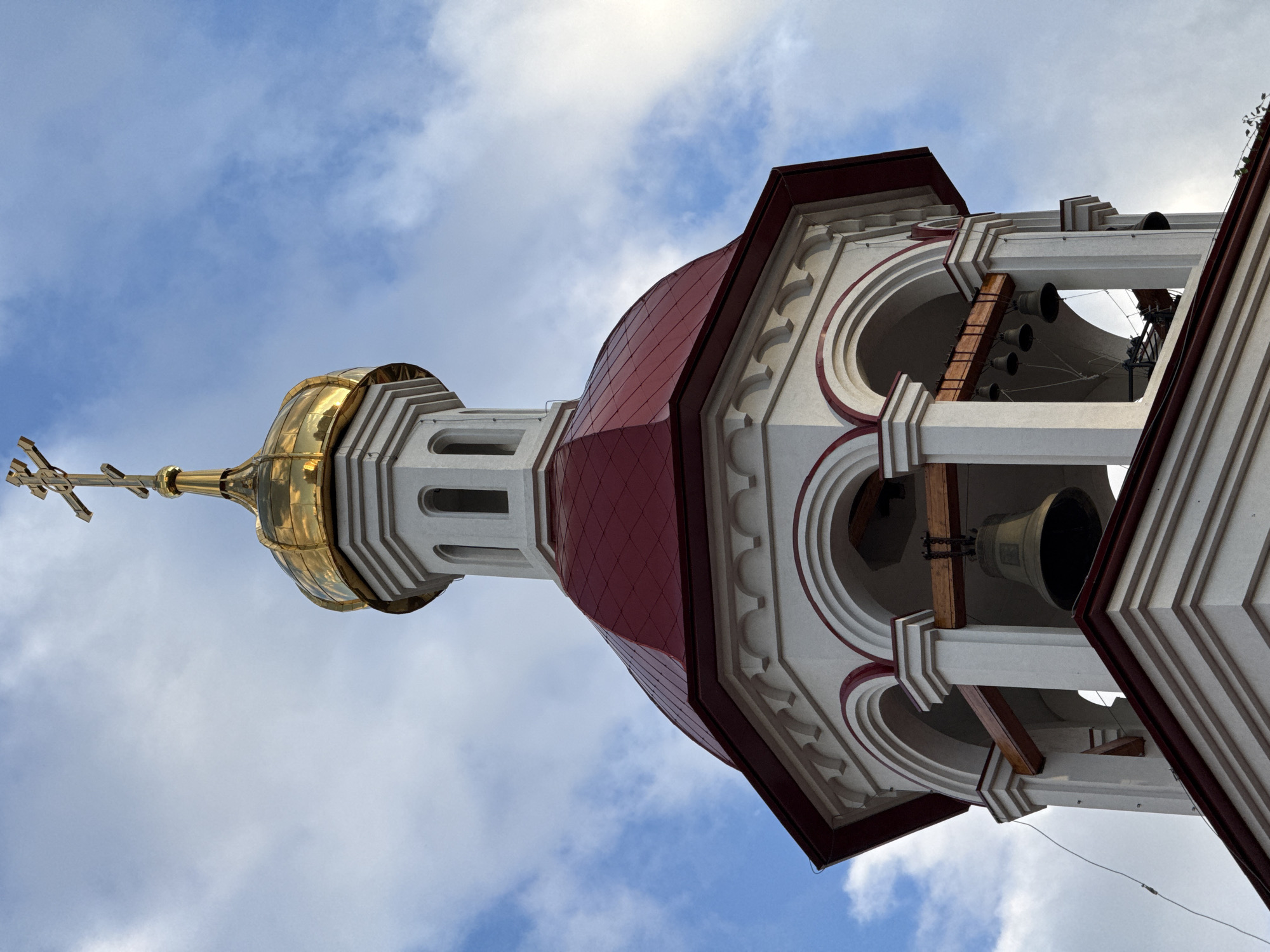 This is a low-angle, rotated shot of a bell tower, likely part of a church, against a cloudy blue sky. The main subject is the ornate architecture of the tower, featuring a golden dome topped with a cross, a red segmented roof, and white and red decorative elements with arched openings. Several large bells are visible hanging within the open sections of the tower. The scene is illuminated by natural light, suggesting it was taken during the daytime. The weather appears to be partly cloudy. No people are visible in the image. The provided location context is Surgut, Russia, which might indicate the architectural style is typical of Russian Orthodox churches. There is no visible text.