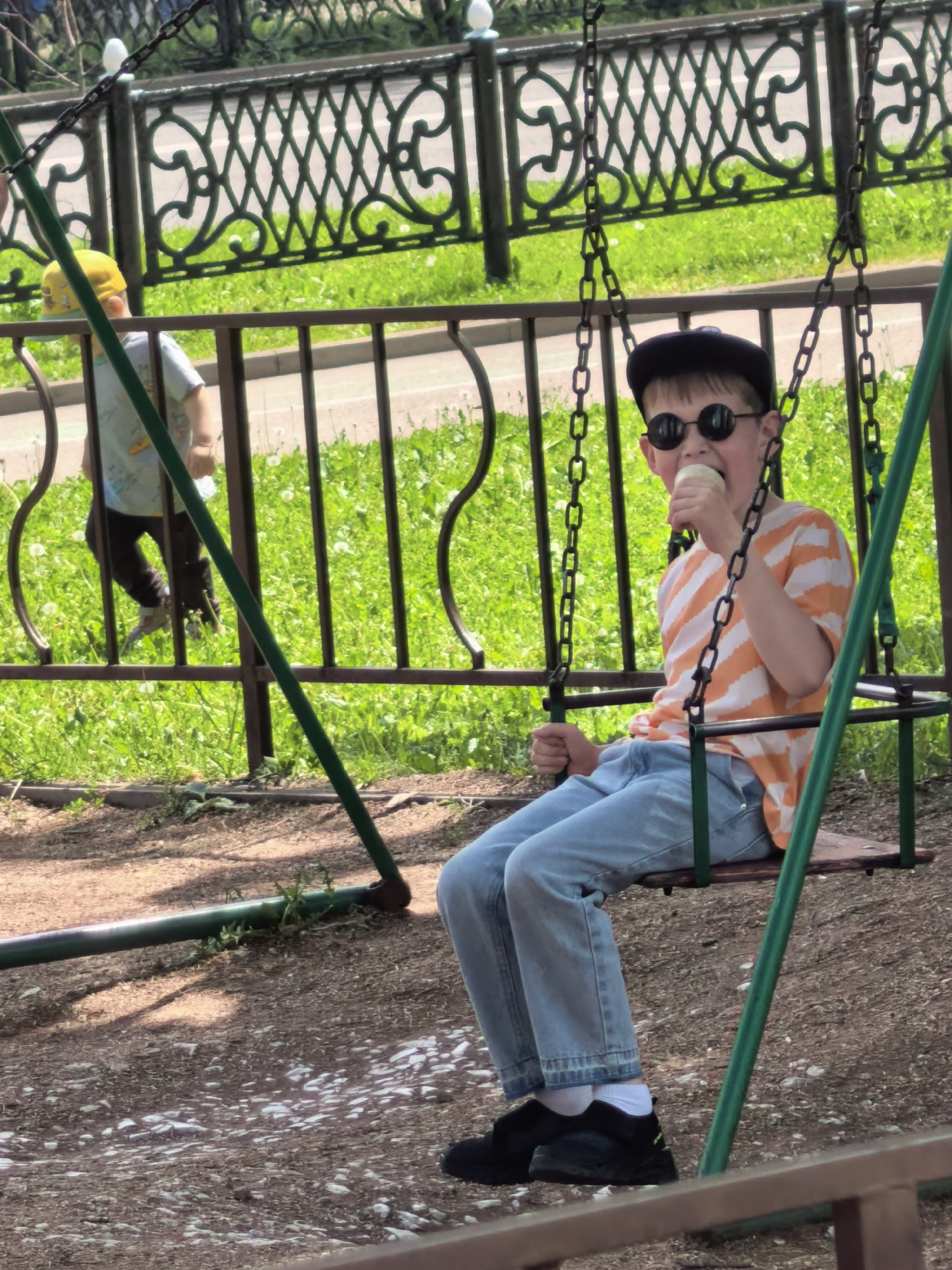 In Sterlitamak, Russia, a young boy sits on a swing, eating an ice cream cone. He wears round sunglasses, a dark cap, and an orange and white striped shirt. His blue jeans are cuffed above black sneakers, revealing white socks. Another child wearing a yellow cap is seen in the background, moving along the fence. The swing set is green, and the area underneath it is dirt and small rocks. A decorative metal fence is in the background. The background also includes grass and other greenery.