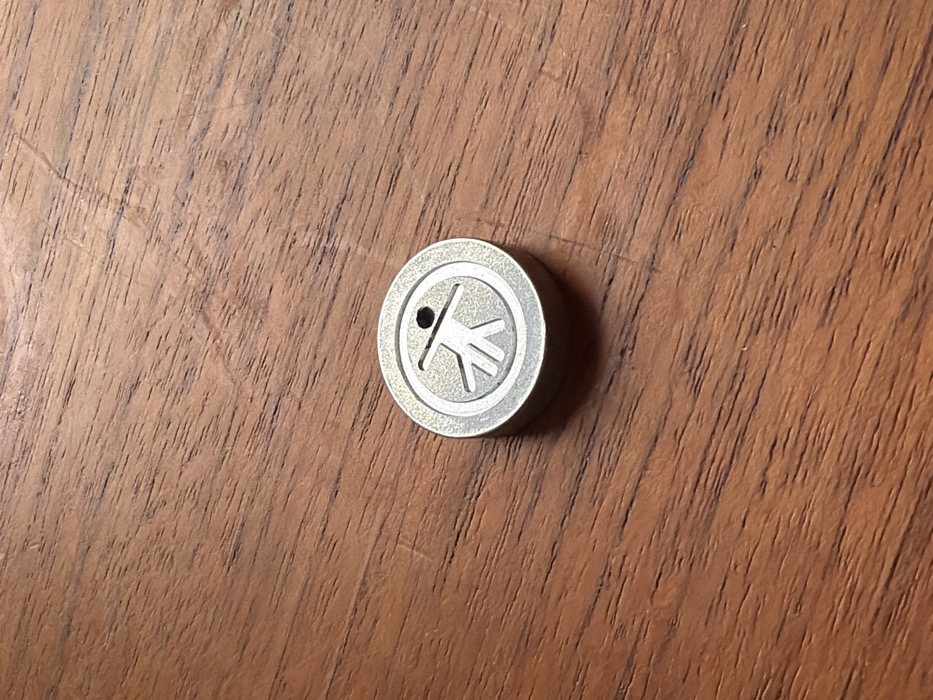 A single, circular, silver-toned metallic token rests off-center on a textured, medium-brown wooden surface. The token's upper face features an etched or recessed symbol: a stylized human figure with splayed legs and outstretched arms, contained within an outer circular border. A small, dark, circular indentation is positioned directly above the figure's head. The wooden background displays prominent grain patterns, minor surface scratches, and variations in tone consistent with natural wood and slight wear. The image contains no visual information, individuals, or contextual cues that allow for the identification of a specific location within the city of London, United Kingdom, or any other geographic area.