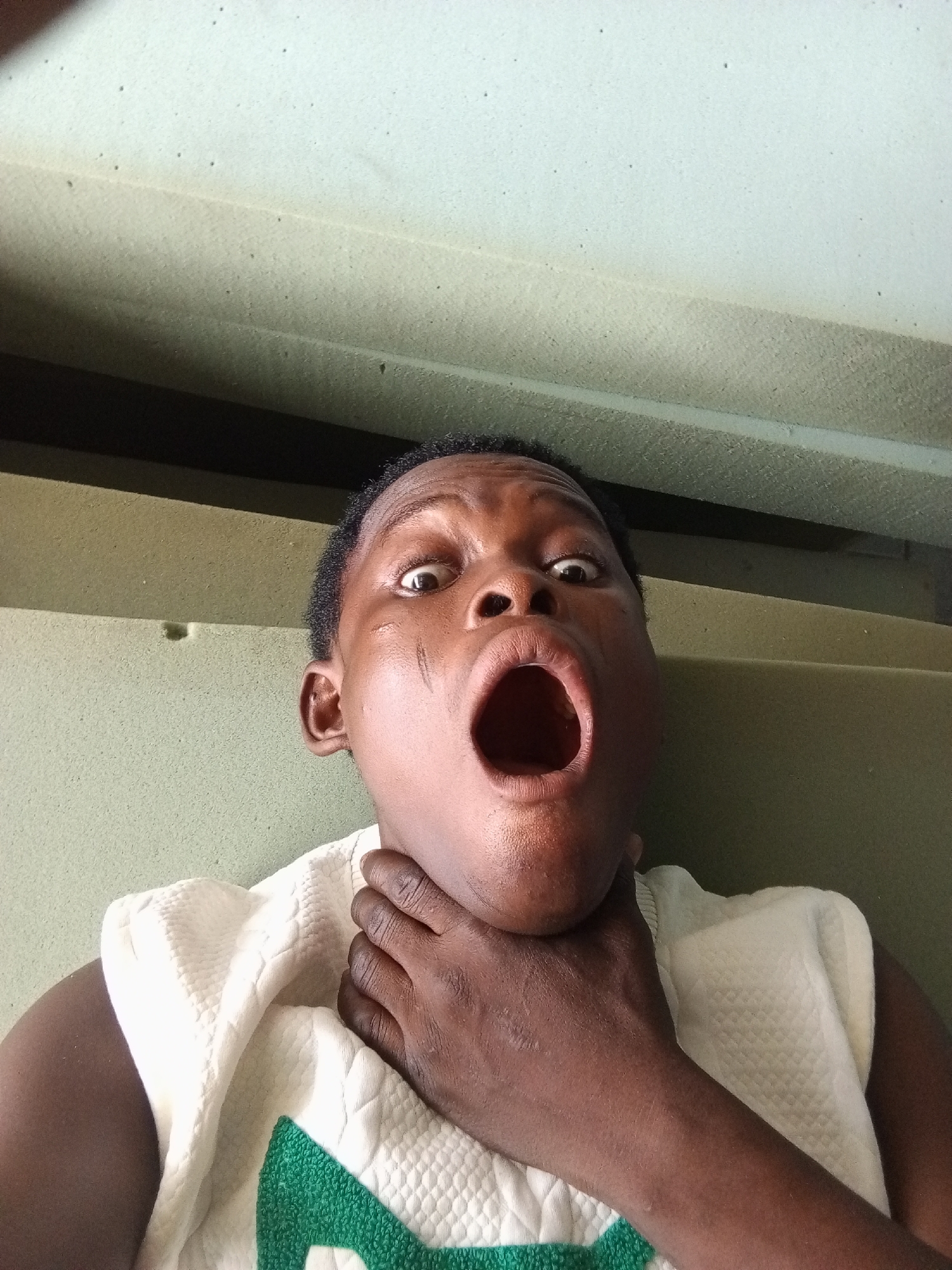 The image captures a close-up, upward-angled portrait of a young individual with dark skin and short, dark hair. The subject is positioned centrally, exhibiting a dramatic expression characterized by widely opened eyes and a large, agape mouth, conveying a sense of shock, surprise, or an exaggerated reaction. Their right hand is placed on their throat, reinforcing the intensity of the portrayed emotion or action.
The person is wearing a light-colored, possibly white, sleeveless top featuring a distinct textured or quilted pattern. A partial glimpse of a green design or logo is visible on the front of the garment. Notable details on the subject's face include faint scratch marks or scars on the left cheek.
The background consists of a textured, light greenish-beige material that resembles foam or upholstered paneling, with darker lines suggesting structural elements or seams. This environment appears to be indoors, potentially a simple room or the interior of a vehicle. Small dark specks are scattered across the background material. The lighting is bright and direct, illuminating the subject's face. No discernible text is visible within the image.