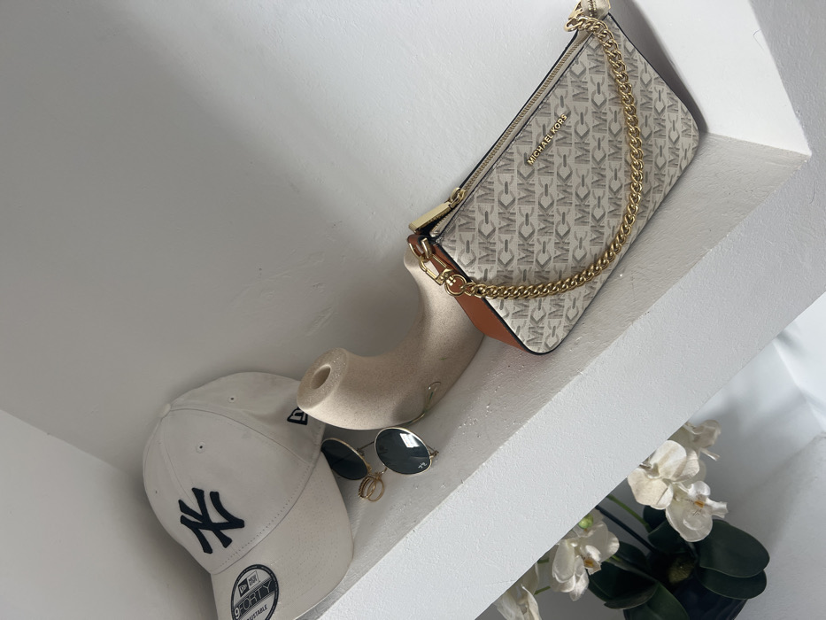 The scene, located in Skhirat, Morocco, depicts several personal accessories arranged on a white, smooth surface, likely a shelf or ledge, with a contrasting diagonal architectural element visible in the lower left. On the left, a white baseball cap, identified by a "NY" logo in dark blue and a "9FORTY ADJUSTABLE" tag, rests. Adjacent to the cap are a pair of gold-framed sunglasses with dark, circular lenses. To their right, a light beige, curved, and hollow object with a circular opening at one end is positioned. On the far right, a Michael Kors brand clutch or small shoulder bag is visible. The bag features a repeating light grey "MK" monogram pattern on a lighter background, brown leather accents on its sides, and a prominent gold-colored chain strap. The "MICHAEL KORS" brand name is visible on the bag's exterior. Partially visible in the bottom-left corner, beneath the white diagonal structure, are a few white orchid flowers with green leaves, emerging from a dark container. No people are present.