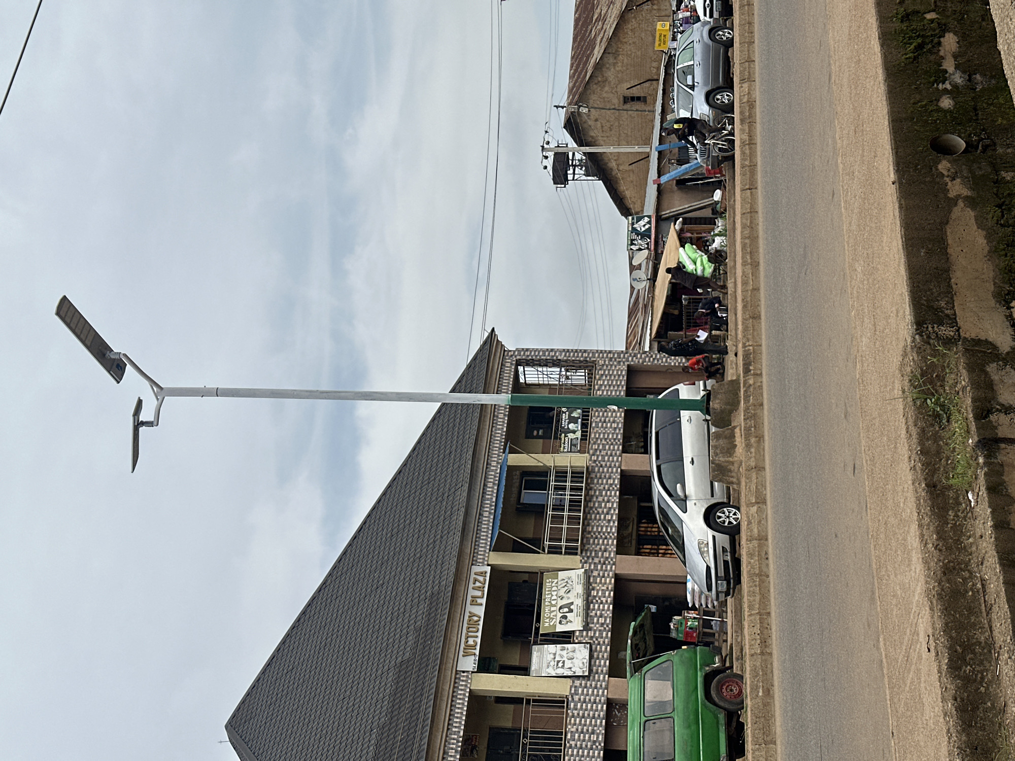 The image depicts a street scene in Bukuru, Nigeria. A prominent building labeled "VICTORY PLAZA" is visible on the left, featuring multiple stories with balconies and various storefronts. In front of the building, a green van is parked, followed by a silver minivan and a silver SUV. Several individuals are present, some standing and interacting near the storefronts and market stalls. A street lamp with solar panels stands tall, illuminating the cloudy sky. In the background, other buildings with corrugated iron roofs and a water tower are visible. The street itself is paved, with a concrete curb and a drainage ditch in the foreground. The overall atmosphere suggests a bustling urban environment.