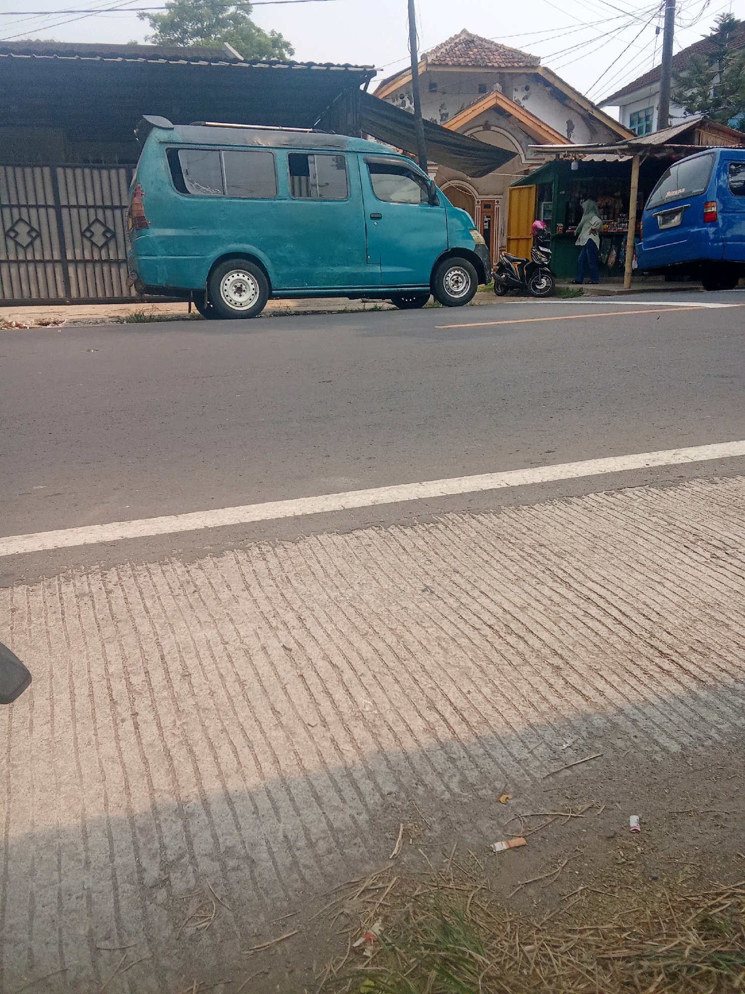 The image captures a street scene in Warunggunung, Indonesia. A teal-colored minivan is parked on the side of an asphalt road, with its rear facing the viewer's left. To the right of the minivan, a small kiosk is set up, and a blue van is parked in front of it. A person wearing a hijab is standing at the kiosk. A black motorcycle with a pink helmet is parked near the kiosk. The environment appears to be a commercial area with buildings and overhead power lines. The foreground shows a textured concrete surface with some scattered debris, including cigarette butts. The lighting suggests it is daytime, possibly late afternoon due to the shadows.