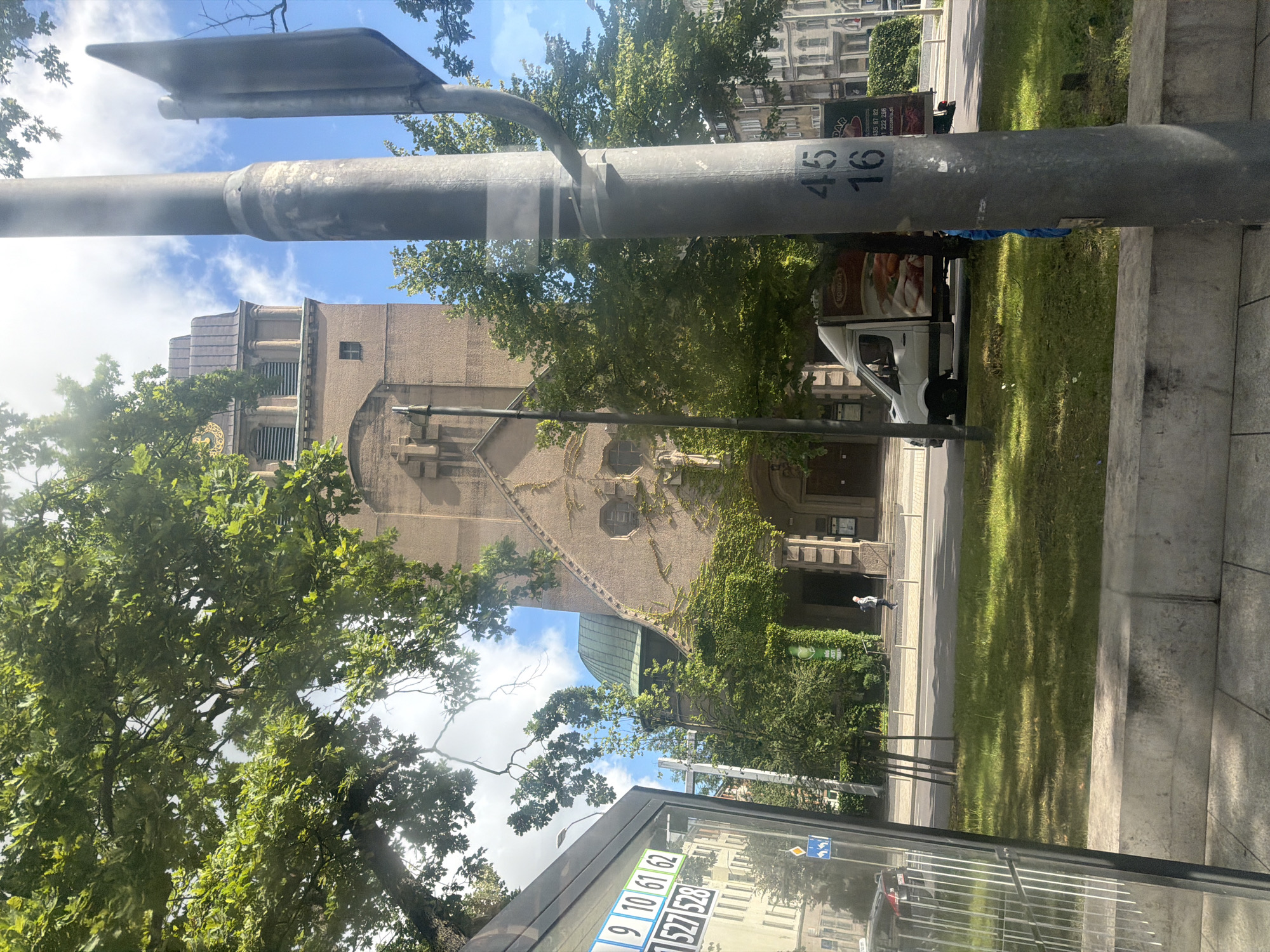 The image captures a street view in Szczecin, Poland, featuring a tall, tan-colored church with a green dome and a prominent cross on its facade. The church is partially obscured by lush green trees, and ivy climbs the building's exterior. In the foreground, a utility pole stands to the right, marked with the numbers "45" and "16." To the left, a bus stop shelter is visible, displaying numbers "9", "10", "61", "62", "527", and "528." A white van with an advertisement featuring food is parked on the street. A single person is walking on the sidewalk in front of the church. The sky is blue with scattered clouds, and sunlight illuminates the scene.