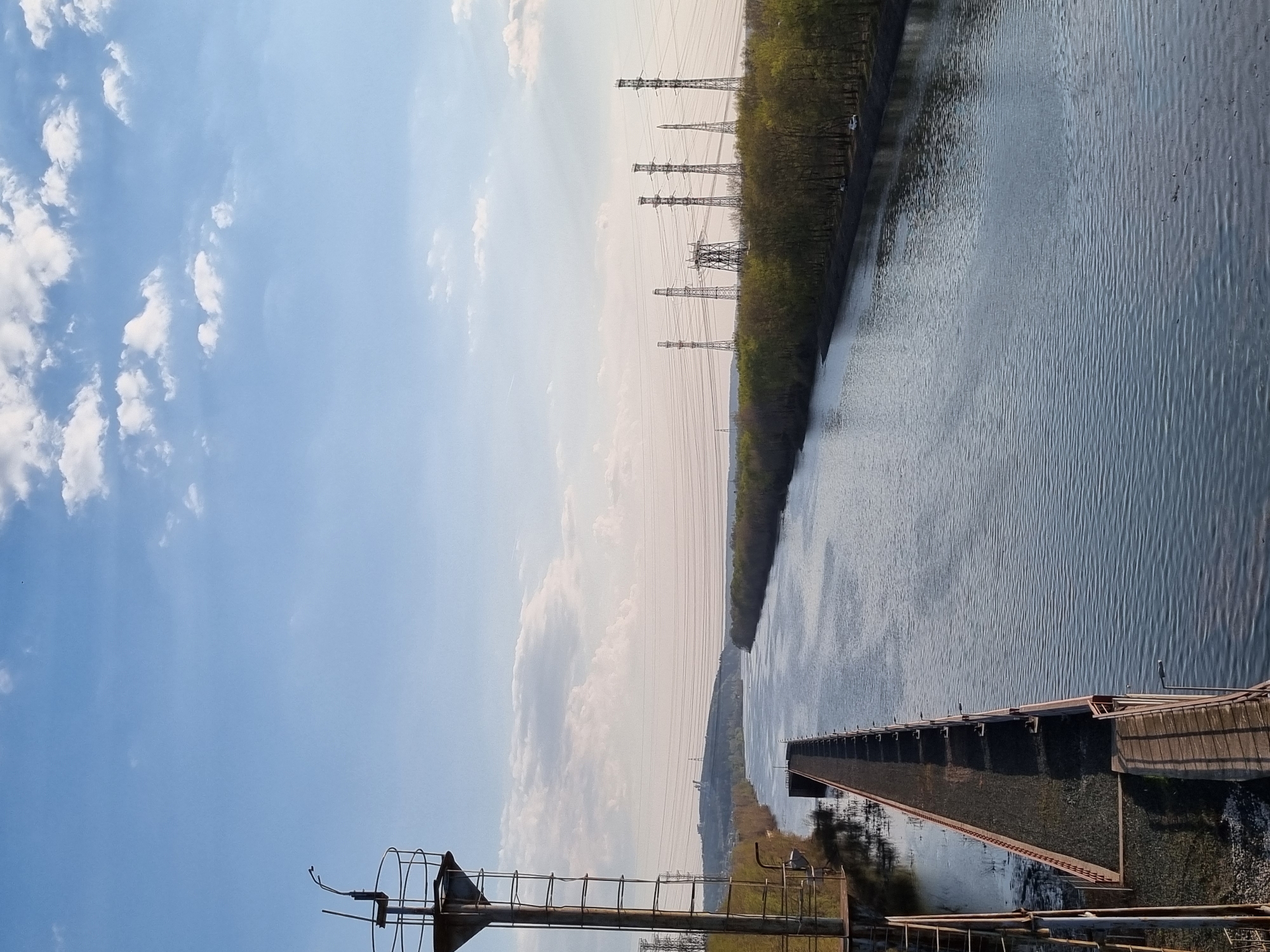 The image shows a canal in the city of Chaikovsky, Russia. The canal, full of water, runs lengthwise through the frame. Its banks are lined with trees and power lines. A bridge structure extends partially across the canal. In the foreground is an industrial structure. The sky is blue with scattered clouds.