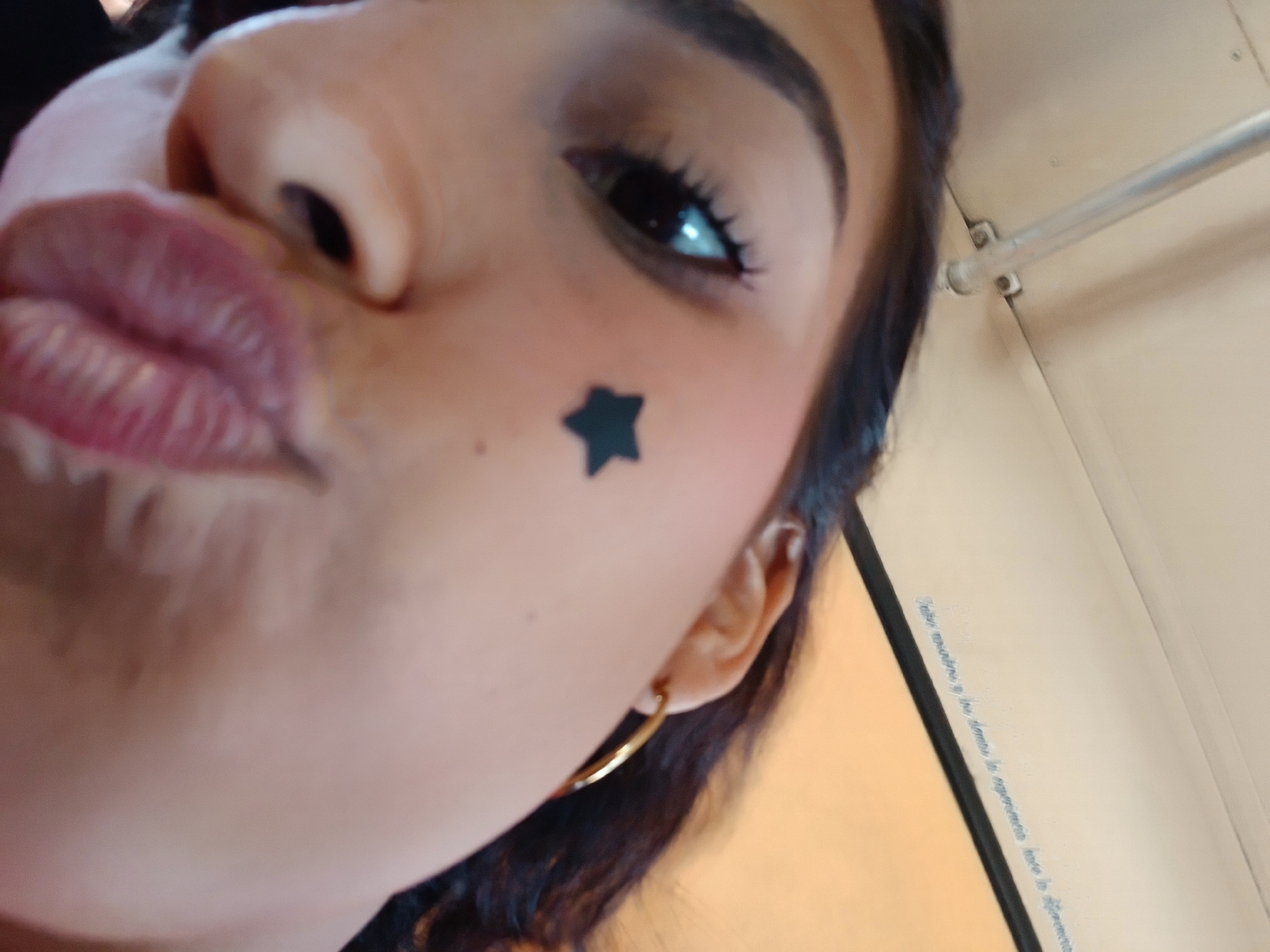 A close-up angled selfie captures a woman with brown hair, light brown skin, and prominent lips puckered in a "duck face." She has a gold hoop earring in her left ear, and a black star is adhered to her left cheek. She is wearing mascara on her upper eyelashes. The background is out of focus, but suggests an interior space with cream-colored walls. There is a metal bar attached to the wall. A barely legible phrase is visible on the wall, written in a small, light-colored font. There is no information to identify this picture as being in Altagracia, Venezuela.