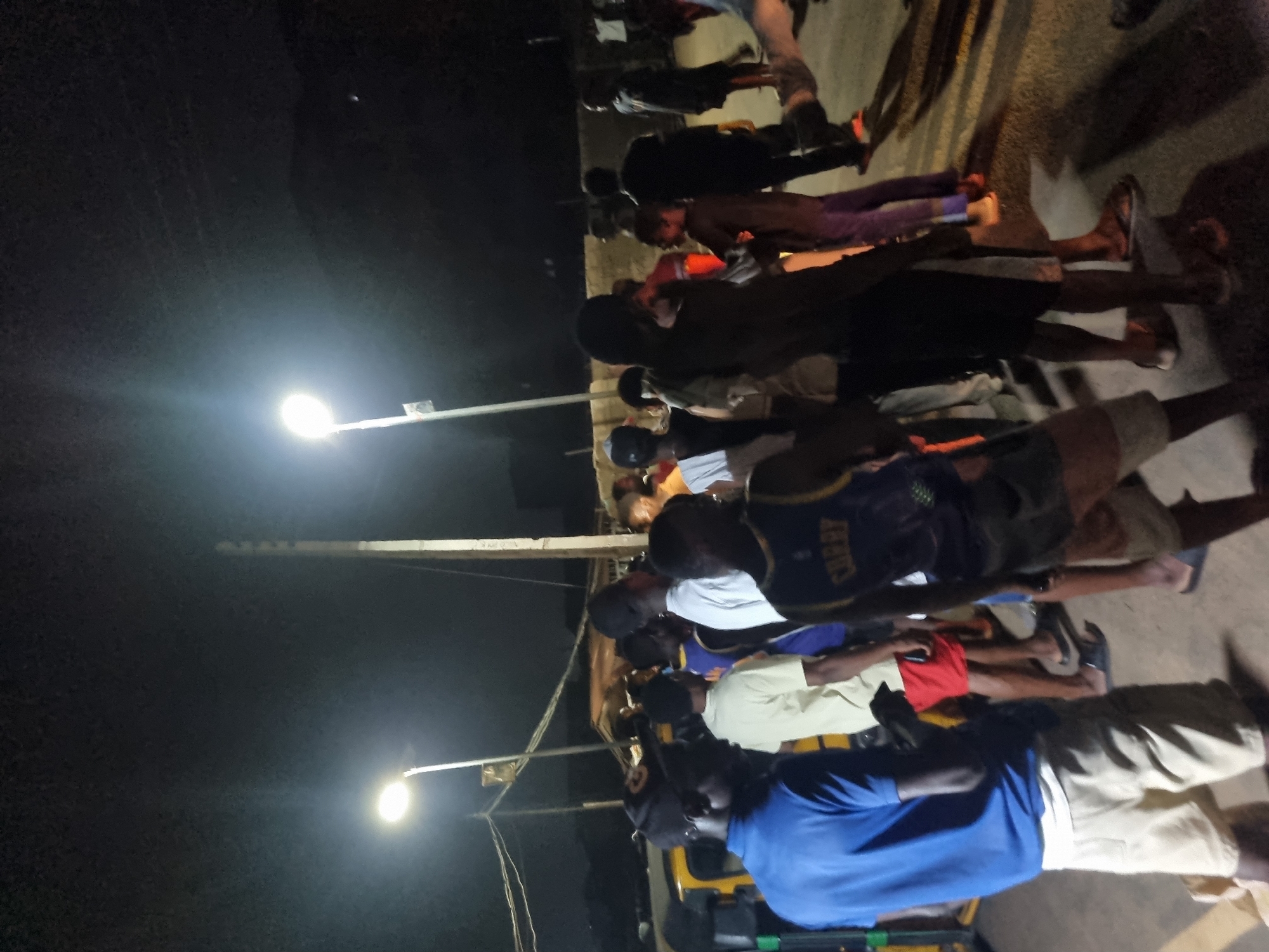 A group of people is standing or walking alongside a street in Onitsha, Nigeria, at night. The scene is illuminated by several street lights. Individuals are wearing a variety of clothing, including shorts, t-shirts, and one person appears to be wearing a basketball jersey with "CURRY" visible on the back. The background includes a partially visible yellow and green vehicle and a dark sky. The ground appears to be paved or concrete.