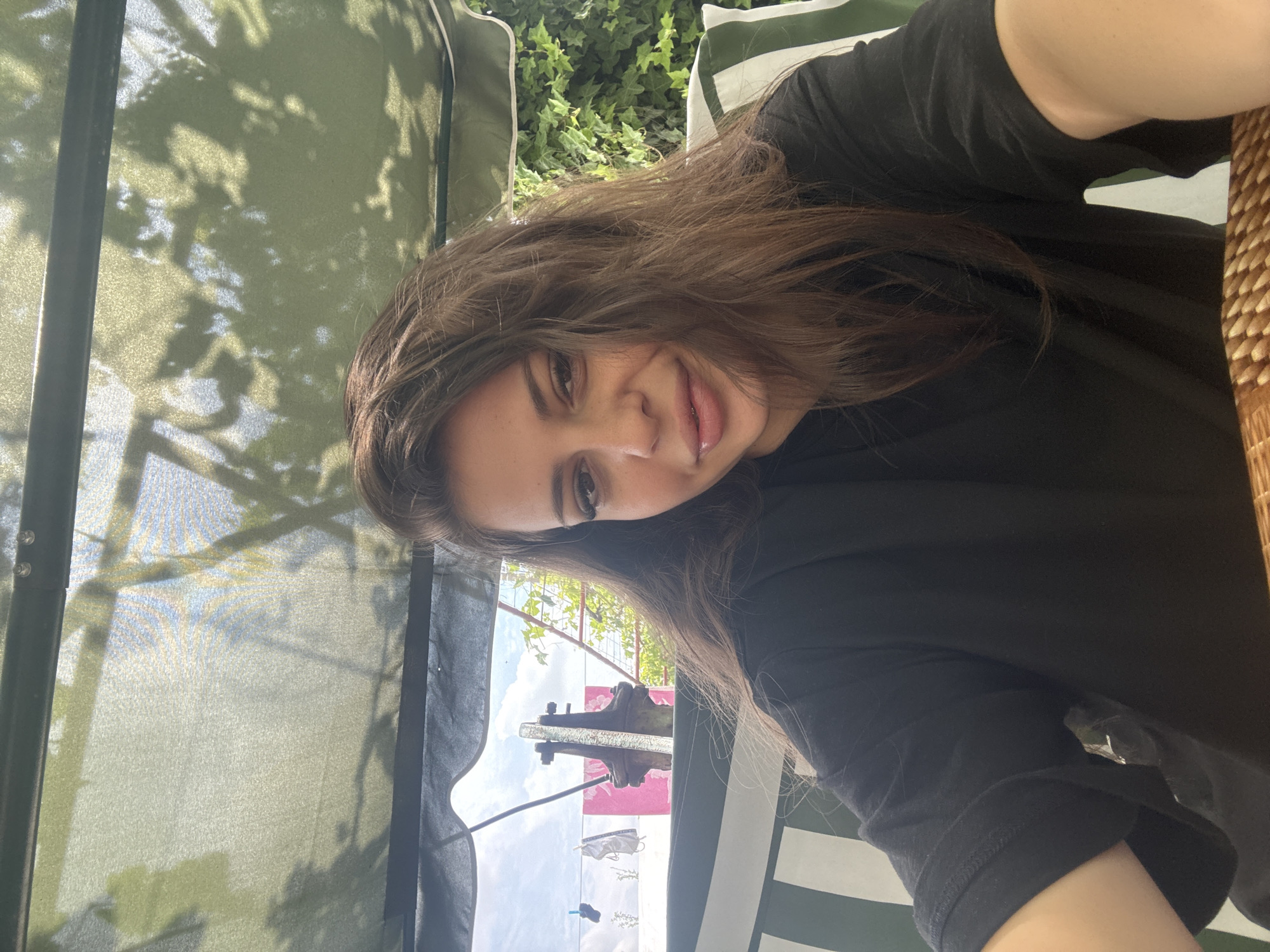 In Govedare, Bulgaria, a young woman with long brown hair, wearing a black t-shirt, is seated on a green and white striped swing. She is looking towards the camera, with her head slightly tilted. The background reveals a green canopy overhead, casting dappled shadows, and a lush green vine wall on the right. In the distance, a clothesline with laundry is visible against a bright sky, along with what appears to be an old water pump.