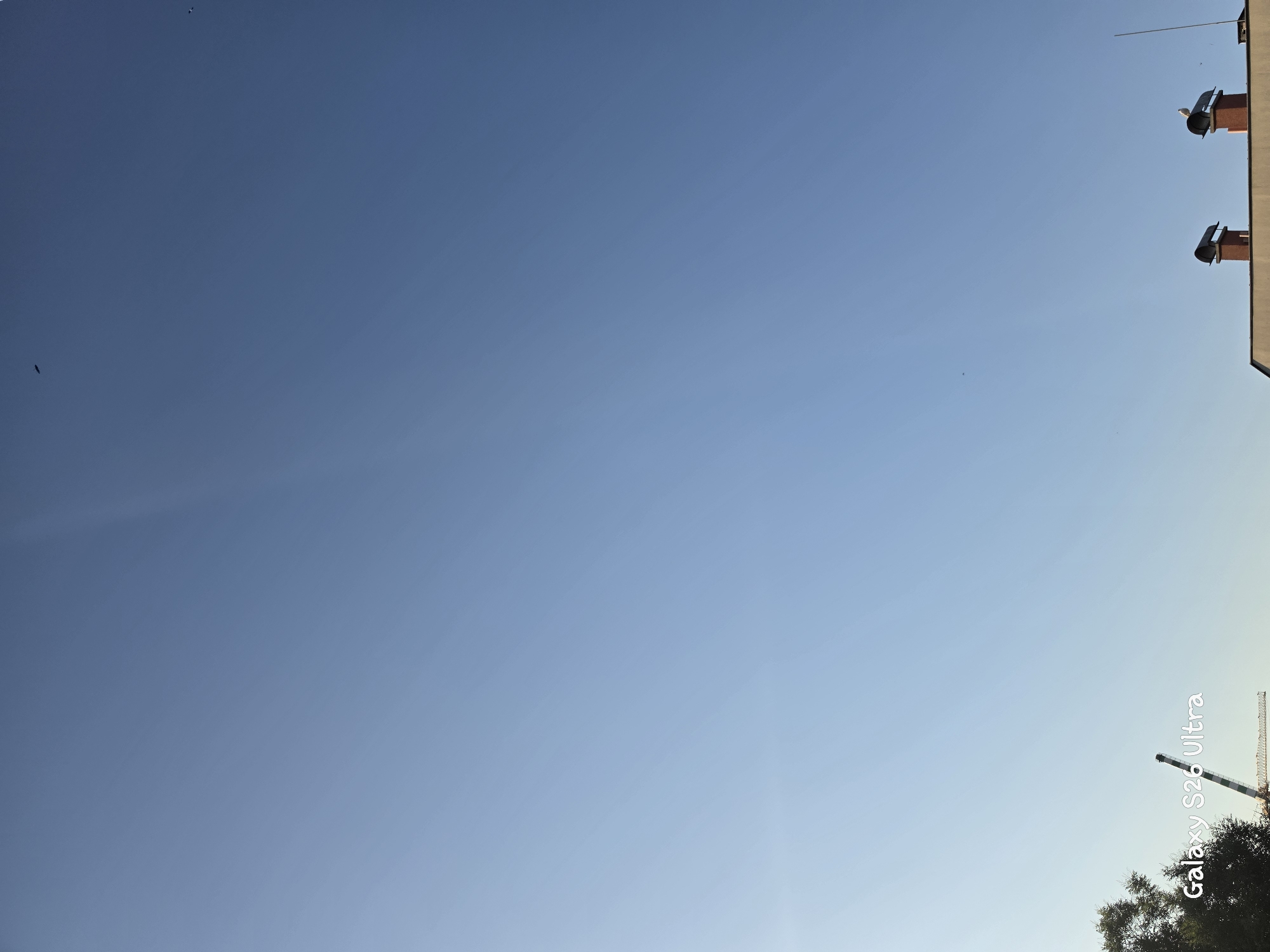 The image is a wide shot of a clear blue sky, with some elements at the bottom and scattered in the sky. In the lower left corner, a portion of a tree is visible, and a construction crane with diagonal yellow and black markings is partially in frame. A watermark reading "Galaxy S26 Ultra" is present on the lower left side. In the lower right, the tops of two brick chimneys with dark caps are visible on a building. A white bird is perched on top of the right chimney. Several small dark specks, likely birds or insects, are scattered across the sky. The scene appears to be an outdoor urban environment on a sunny day.