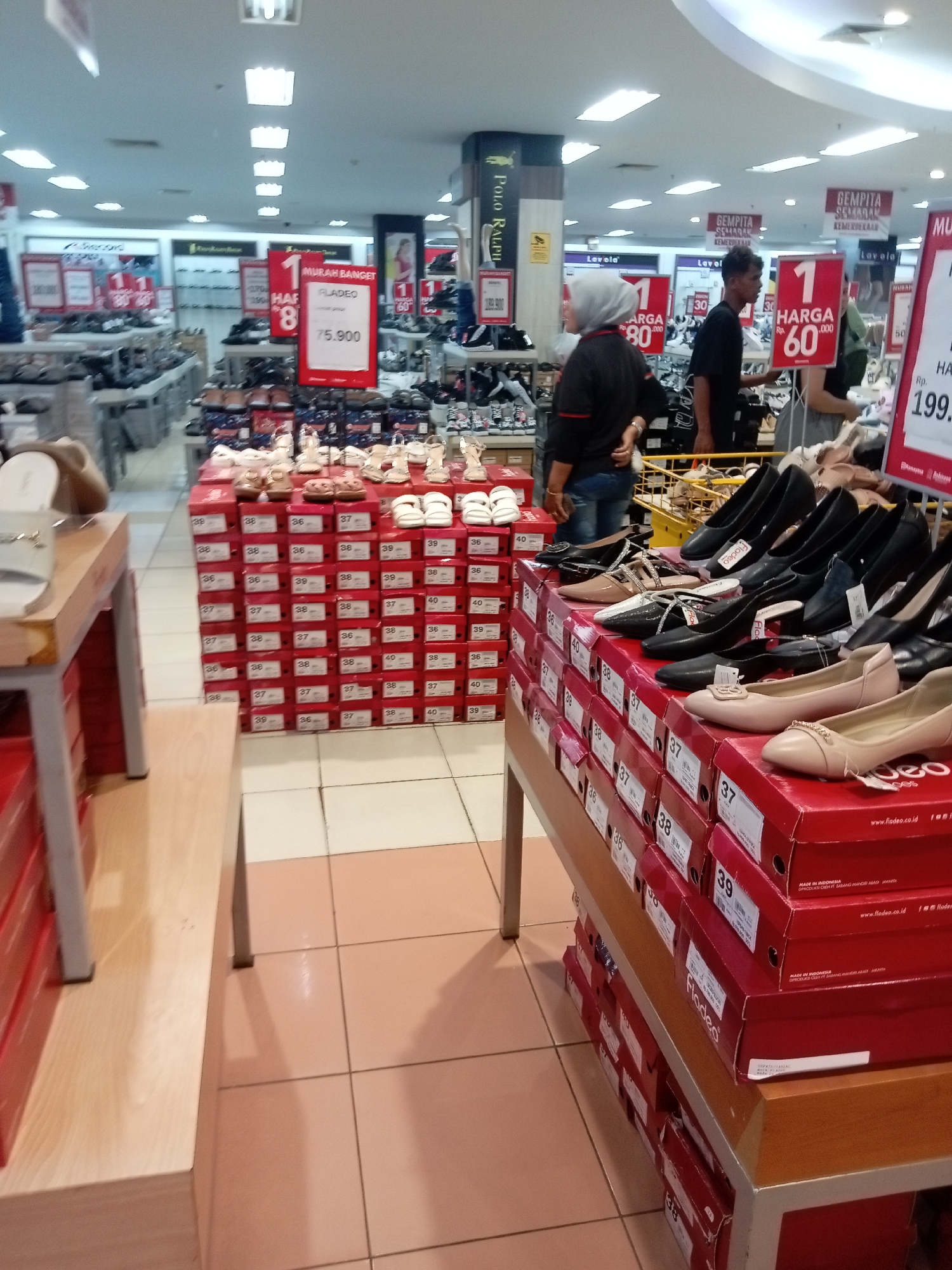 The image depicts the interior of a shoe store, likely located in Batam, Indonesia, given the presence of Indonesian text on sale signs and product boxes. The store features extensive displays of women's footwear, organized in red boxes stacked on shelves and on top of tables. Several pairs of high-heeled shoes and flat shoes are displayed on top of the red boxes. In the background, rows of other shoes are visible on display racks.
The scene shows several individuals, including a woman wearing a grey hijab and a dark top with red accents, and a man in a dark t-shirt and jeans. They appear to be browsing or working in the store. The environment is brightly lit by overhead fluorescent lights. Price tags and promotional signs are visible, indicating a sale with prices such as "Rp. 75.900" and "Rp. 199.900". The flooring is tiled with light-colored squares. The overall impression is a retail space designed for the sale of footwear.