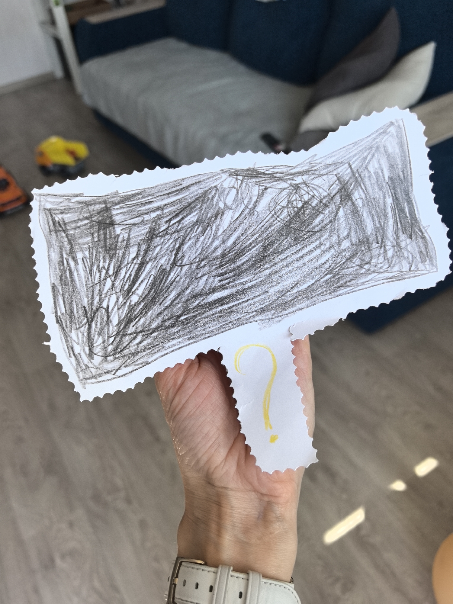 A person holds a white, rectangular piece of paper with a zig-zag cut edge. The paper has a drawing in dark pencil or crayon covering most of the rectangular area. Below the rectangle, a smaller piece of white paper, also with a zig-zag edge, is attached, bearing a yellow question mark. The person holding the paper is wearing a white watch with a white leather strap and silver buckle. The background is out of focus but includes a wooden floor, a blue couch with light grey and dark grey pillows, and some toys. The image does not appear to depict any specific location in the city of Perm, Russia, and the lack of detail or context in the image prevents this.