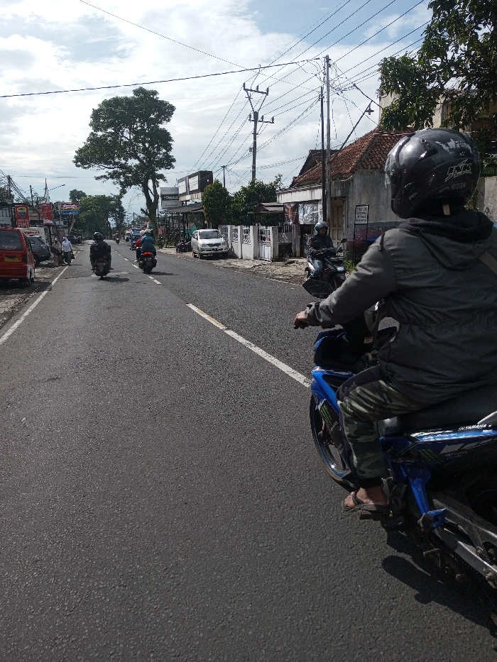 The scene depicts a daytime street in Domphoog_Dungkarchhoeling, Bhutan, viewed from the perspective of a person on a motorcycle. The asphalt road features a solid white lane divider that transitions to a broken line further down.
In the immediate foreground, facing away from the viewer, a person is seated on a blue Yamaha motorcycle. This individual wears a black full-face helmet with white markings, a dark grey hooded jacket, camouflage-patterned trousers, and dark sandals. The motorcycle exhibits blue bodywork with dark green/black graphic decals. A shadow from the foreground motorcycle is cast onto the road surface.
Ahead, three other motorcycles are visible, traveling away from the camera in the same direction. Their riders are also helmeted and clad in dark clothing. On the far left of the road, a red minivan or small car is parked or moving slowly, followed by additional parked vehicles and at least one pedestrian, dressed in white, walking on a sidewalk or shoulder.
The background along the left side of the street features multiple buildings, some displaying commercial signage with partially visible text, including "RKI." A large, mature tree with dense green foliage stands prominently on this side. Along the right side of the street, a white building with a reddish-brown tiled roof is visible, along with white fences featuring decorative metalwork. Numerous utility poles with a complex network of overhead power lines crisscross the sky, particularly on the right side. The sky is overcast with significant cloud cover but indicates a bright daytime setting.
