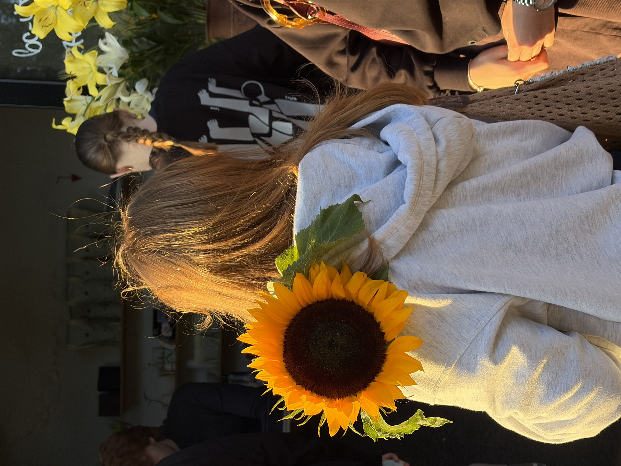 The image shows a close-up of two individuals from the shoulders up, with a sunflower prominently placed in the foreground. The person in the foreground has long, auburn hair that is illuminated by golden sunlight, suggesting it is either sunrise or sunset. They are wearing a light gray hooded sweatshirt. A single sunflower with bright yellow petals and a dark center is positioned on their shoulder, with its stem and leaves visible.
The person behind them is wearing a black hooded sweatshirt with a graphic design featuring stylized human figures. Their hands are clasped together in the foreground, with one wearing a watch and a bracelet. The background is blurred but suggests an outdoor setting, possibly a cafe or seating area with woven furniture. The overall lighting is warm and soft, casting long shadows. The presence of a sunflower and the warm light imply a pleasant, possibly relaxed atmosphere. The image is cropped tightly, focusing on the interaction and the sunflower. The context provided indicates the location is Minsk, Belarus.