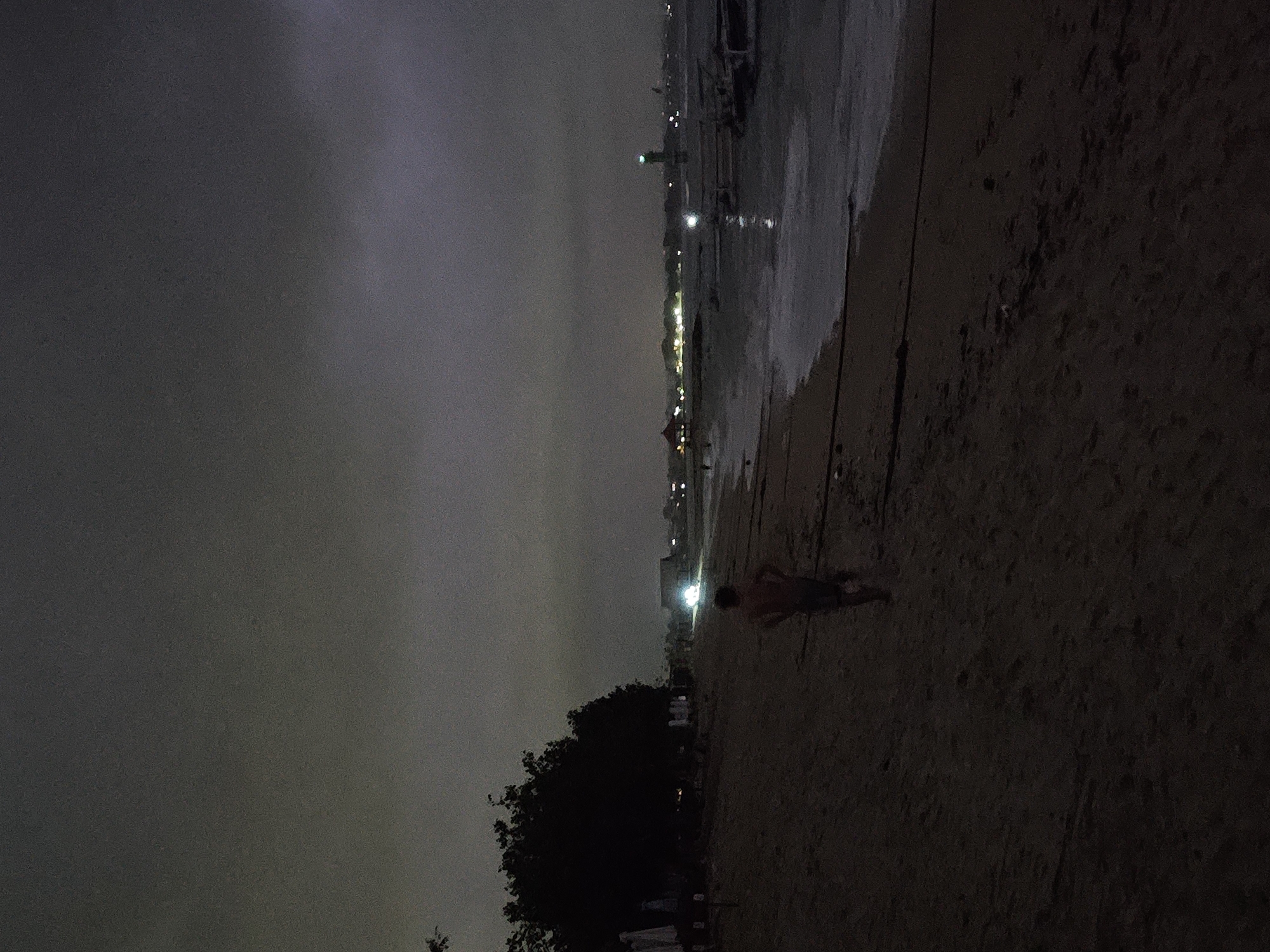 The image depicts a beach at night in Denpasar, Indonesia. The sky is overcast. The shoreline is visible, with waves breaking. A person stands on the sand near the water. In the distance, city lights are visible along the coast. Trees are present near the shoreline.