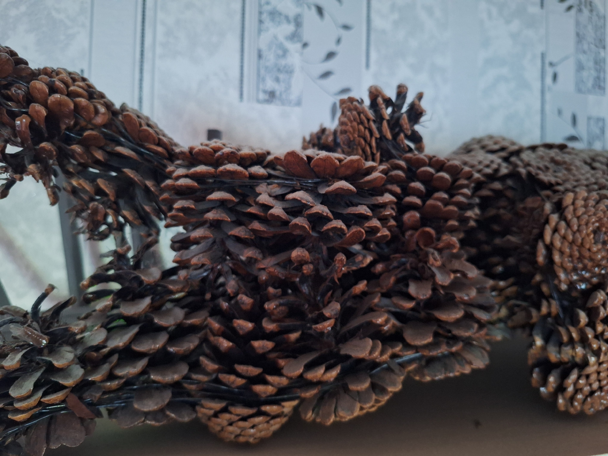 The image shows a close-up, low-angle shot of a decoration made of pine cones. The pine cones are dark brown and appear to be attached to a wire frame. They are arranged in clusters and rows, creating a textured, organic surface. The background is a blurry light blue and white pattern, possibly wallpaper or a textured surface. The lighting is soft, highlighting the details of the pine cones. There are no people or animals visible in the image. The scene suggests an indoor setting, likely a home or a craft fair, given the decorative nature of the pine cone arrangement. The location in Varna, Bulgaria, cannot be determined from the visual information provided in the image.