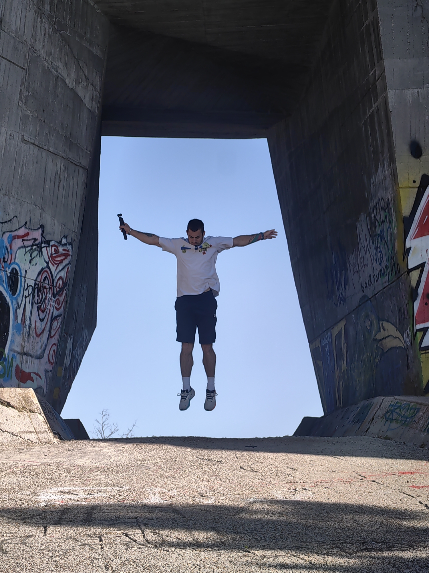A man is jumping in the air, with his arms outstretched and holding a selfie stick in his right hand. He is wearing a white t-shirt, black shorts, and gray sneakers. The man is positioned within a concrete structure that is likely a bridge or underpass. The walls of the structure are covered in graffiti. The sky is visible through the opening of the structure, and there is a paved surface below. The image was taken in Varna, Bulgaria.
