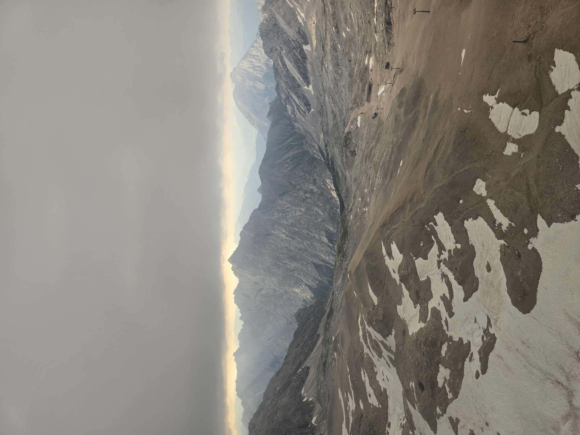 The image depicts a vast, barren mountainous landscape under a cloudy sky. In the foreground, the ground is a mixture of dry, rocky earth and patches of dirty snow. The terrain slopes gently upwards towards a range of rugged, grey mountains that dominate the middle ground. These mountains are heavily textured, with sharp peaks and deep valleys, some of which are shrouded in mist.
Further in the distance, more mountain ranges are visible, fading into a hazy, atmospheric perspective. The sky is overcast with heavy, grey clouds that obscure the sun, but a faint glow of light breaks through in the horizon, illuminating the distant peaks with a soft, golden hue.
In the middle ground, on a relatively flat section of the mountainside, there are signs of human infrastructure. A few ski lift pylons are visible, along with what appears to be a small building or station. There are also a few scattered figures, too small to discern details, walking on the dusty ground. The overall impression is one of a remote, high-altitude environment, likely a ski resort or a hiking area, during a period of inclement weather. The location is the Zugspitze, the highest peak in Germany, near Garmisch-Partenkirchen.