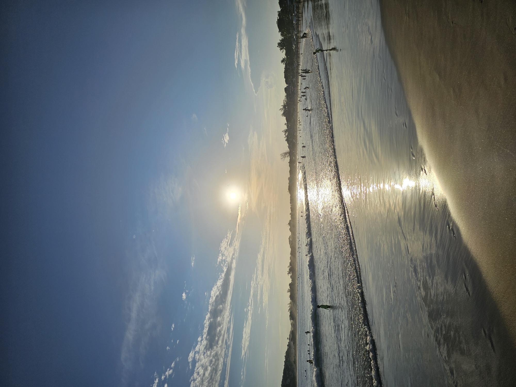 The image captures a beach scene in Armação dos Búzios, Brazil, likely during sunset. The sun is positioned near the horizon, partially obscured by clouds, casting a bright glare across the water's surface. The sky is predominantly blue, with wisps of white clouds scattered. Along the shoreline, people are visible, some standing on the sand, while others are wading in the water. The beach itself appears to be sandy, with a mix of wet and dry areas, reflecting the sunlight. The shoreline is lined with trees and buildings.