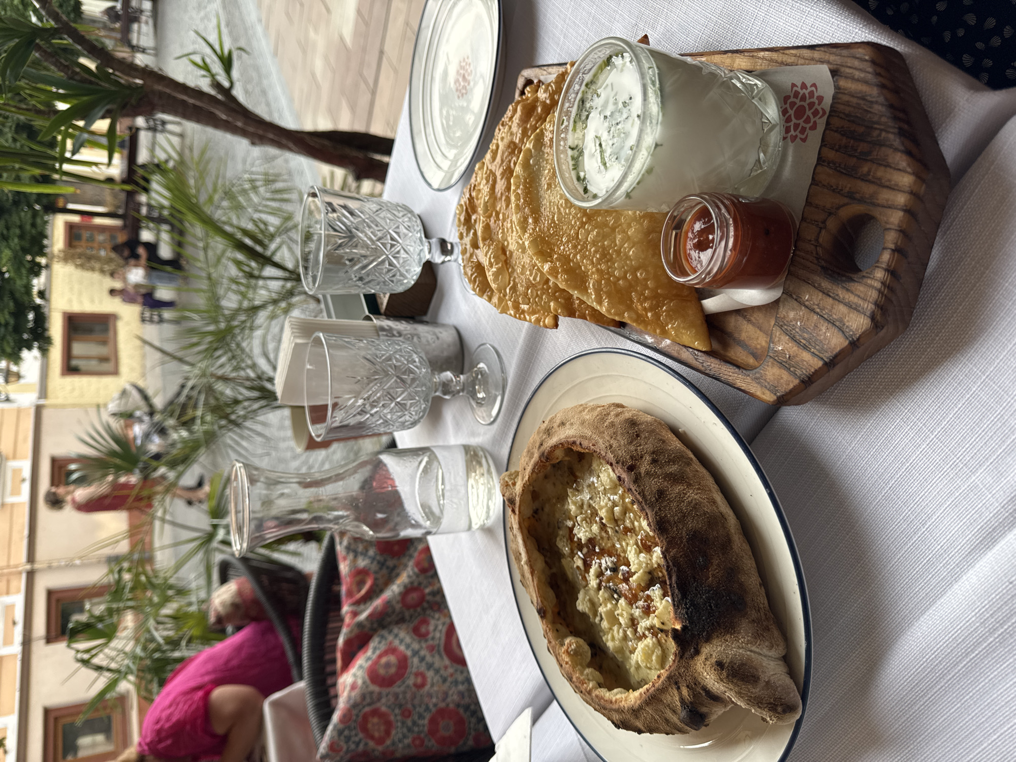 A table set for a meal is shown outdoors, with a white tablecloth, cut crystal glasses, and a large, boat-shaped bread filled with cheese and sauce. A wooden board holds a jar of white dip with herbs, small jars of red sauce, and fried pastries. In the background, people are visible on a street with buildings, and greenery from trees and plants adds to the setting. The overall scene suggests a casual dining experience on a sunny day, possibly in a European city given the architecture. The focus is on the food and table setting, with the background providing a glimpse of the surroundings.