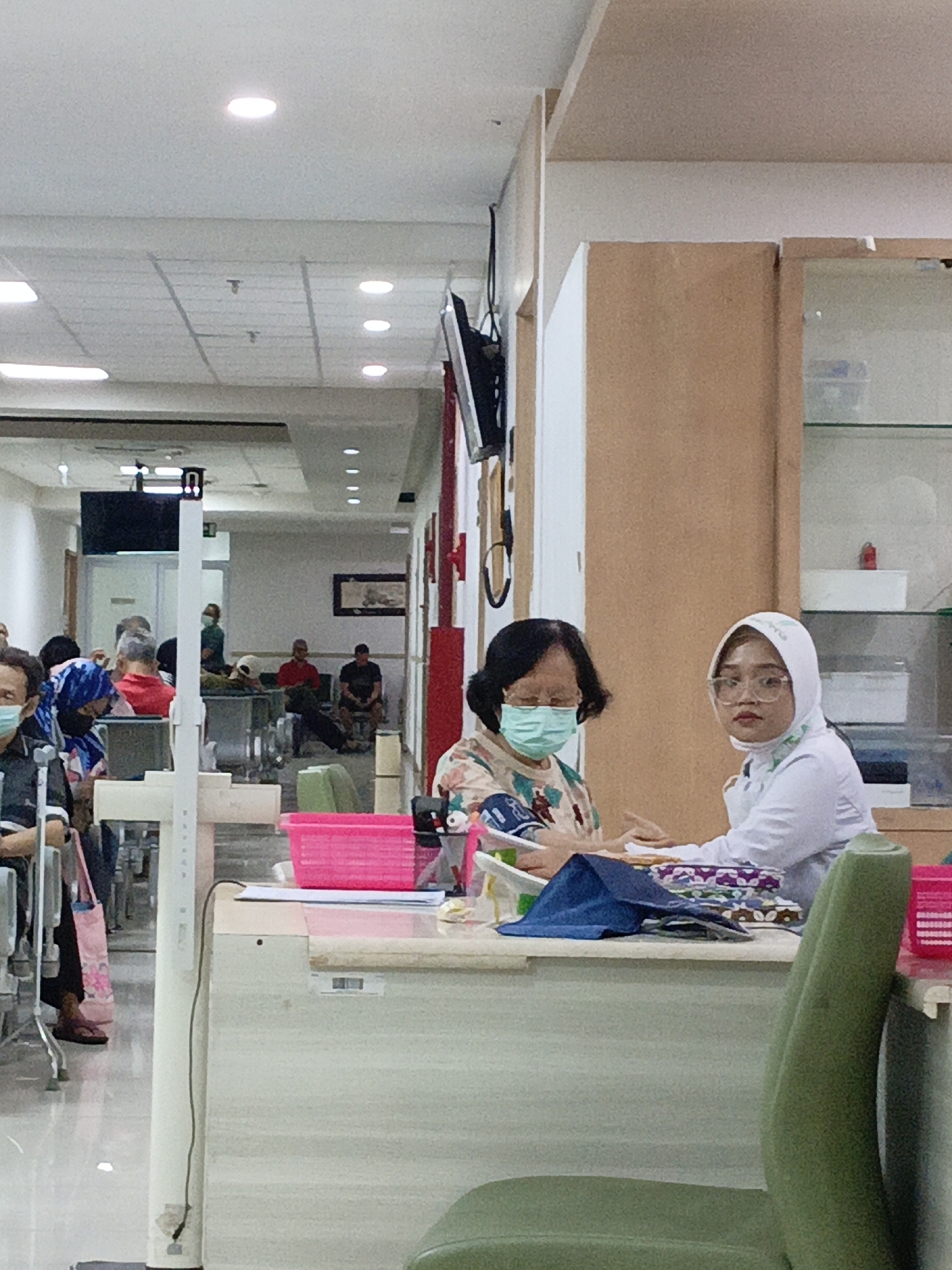 The image depicts a healthcare setting, likely a clinic or hospital waiting area, in East Jakarta, Indonesia. In the foreground, a patient wearing a light-colored floral blouse and a light blue mask is being attended to by a healthcare professional. The professional, wearing a white hijab and glasses, is dressed in a white uniform and appears to be taking the patient's blood pressure. They are seated behind a counter or desk, on which various medical supplies and personal items are present, including a pink plastic bin.
The background reveals a waiting area populated by several other individuals, some seated, some standing, and all appearing to be waiting for medical attention. The environment is brightly lit with fluorescent ceiling lights. The overall impression is one of a typical urban healthcare facility in Indonesia.