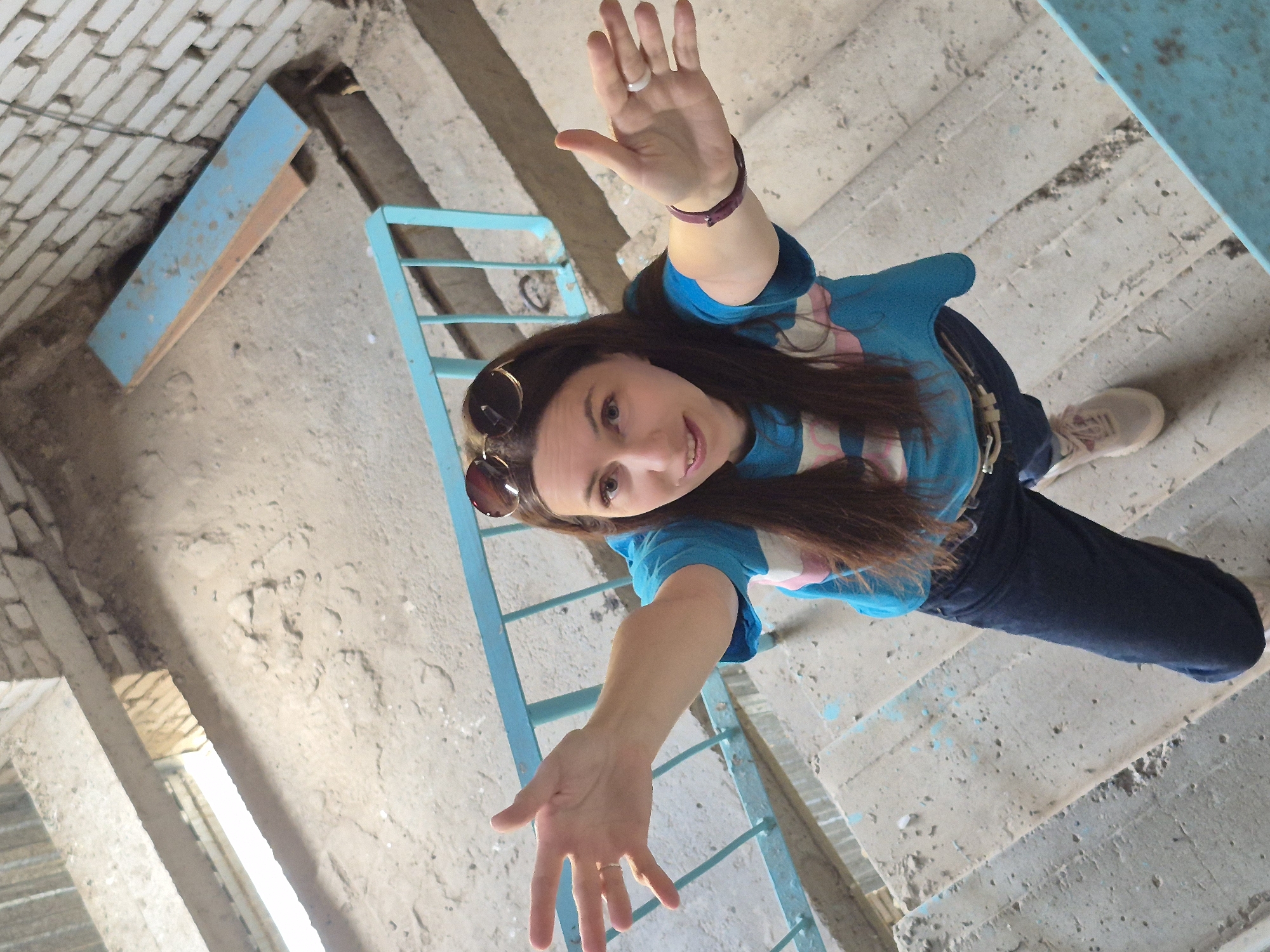 A woman with long brown hair, wearing sunglasses, a blue and pink striped shirt, and dark jeans, stands on a concrete staircase. She is looking up at the camera with her arms outstretched, palms facing outwards. A light blue metal railing is visible on her right. The staircase and surrounding area appear to be an outdoor or semi-outdoor structure, possibly part of an apartment building in Chaikovsky, Russia. The environment is somewhat dilapidated, with visible wear and tear on the concrete and some debris.