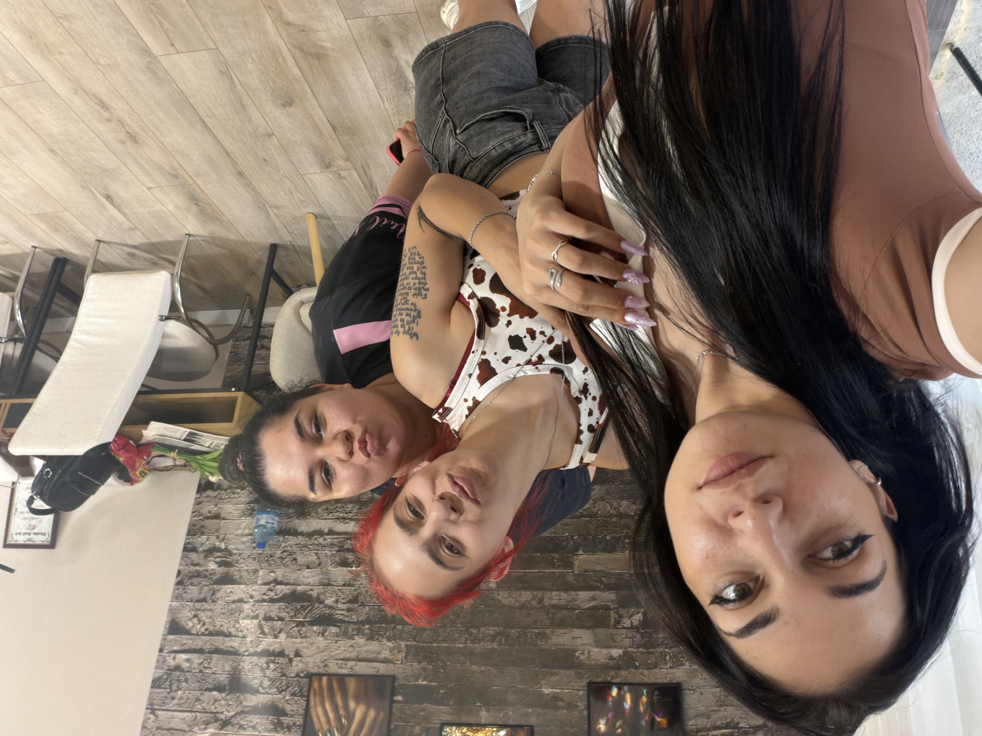 This indoor photograph, taken in Stara Zagora, Bulgaria, depicts three women posing for a selfie.
From left to right:
The first woman, closest to the camera, has long, straight dark hair, parted slightly to her left. She wears a light brown top with a white trim and a silver chain necklace. Her left ear features a silver hoop earring, and her eyes are accentuated with black winged eyeliner. Her nails are long, manicured, and appear to be light pink or lavender with a glitter finish. She looks directly into the camera with a slight smile. Her right hand is interlocked with the left hand of the second woman.
The second woman, positioned behind and slightly to the right of the first, has bright red, medium-length hair. She wears a white tank top with a brown cow-print pattern, a silver chain necklace, and several silver rings on her fingers. A silver braided bracelet is visible on her right wrist, and text-based tattoo art is present on her upper right arm. She is looking at the camera with a slight pout.
The third woman, furthest back and to the right, has dark hair tied up in a bun. She wears a black top with pink detailing on the shoulder/sleeve. Her head is tilted, and she presents a pouting expression towards the camera. A red object, possibly a bracelet or watch, is visible on her left wrist.
The background features a textured wall composed of grey and brown stone-like panels. Three framed art pieces are mounted on this wall; the central one displays abstract colorful elements, and the rightmost one shows a textured brown object. To the right, a light-colored wooden desk or table is visible, holding a plastic bottle of water, a small bouquet of white flowers wrapped in paper, various papers, a red and yellow snack bag, a small black purse, and a framed "Studio Nail Art Certificate." Two white chairs with silver frames are partially visible around the table. The floor is covered with light-toned wooden laminate.