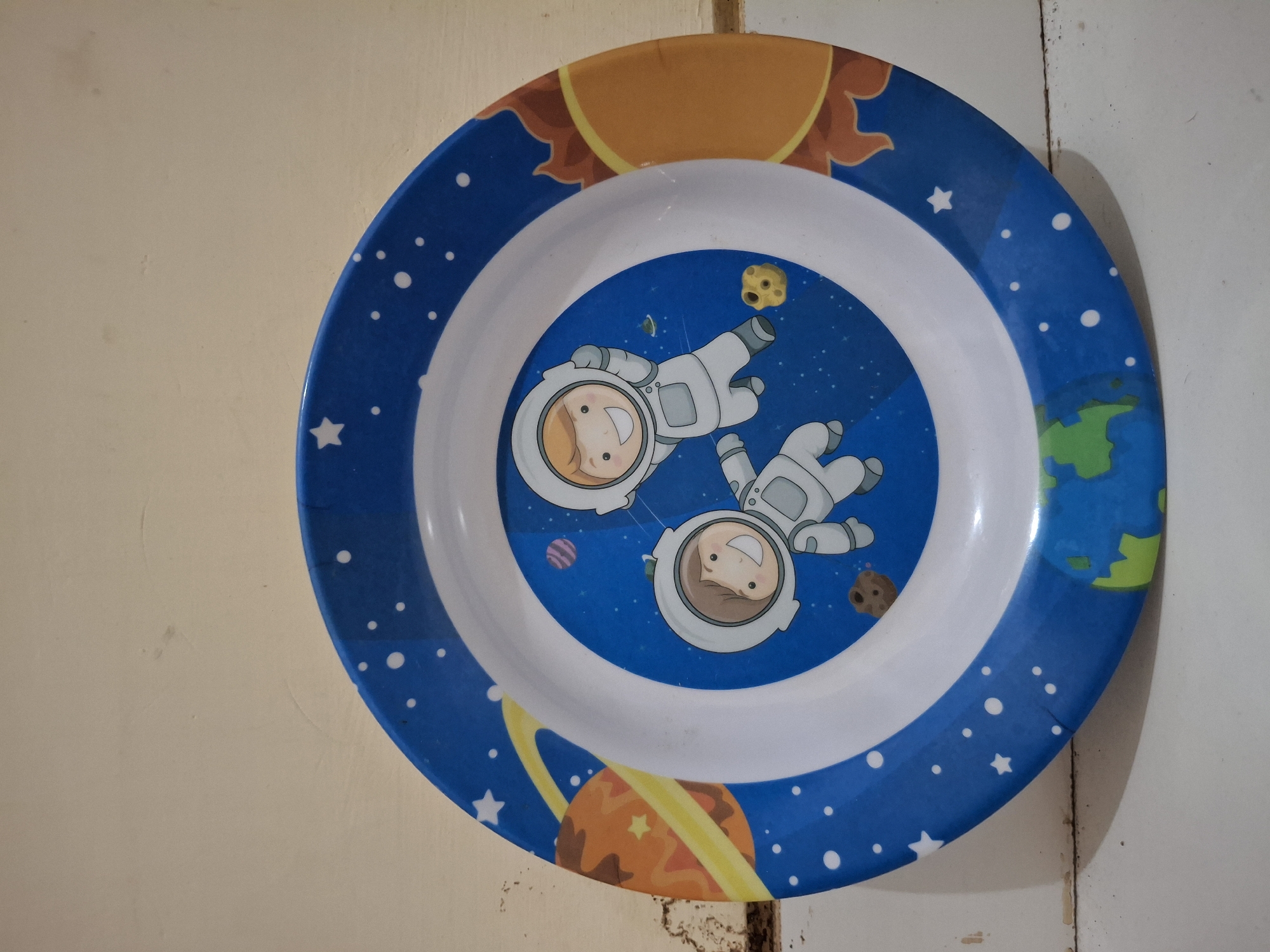 An eye-level, medium shot captures a circular, plastic plate with a space-themed design, set against a beige wall and a white surface. The plate features a vibrant blue background sprinkled with white stars, depicting two cartoon astronauts in white suits floating in outer space. One astronaut is positioned higher than the other, and both have friendly smiles. Surrounding them are illustrations of planets, including a ringed orange planet to the left and a representation of Earth at the bottom. A bright yellow and orange sun with flame-like rays is visible on the right side of the plate. The plate has a raised white rim, and the overall design suggests it is intended for children. The background shows a textured beige wall with faint marks and a white surface, possibly a table or floor, with some debris. The scene is devoid of any explicit indication of location in Banguntapan, Indonesia.