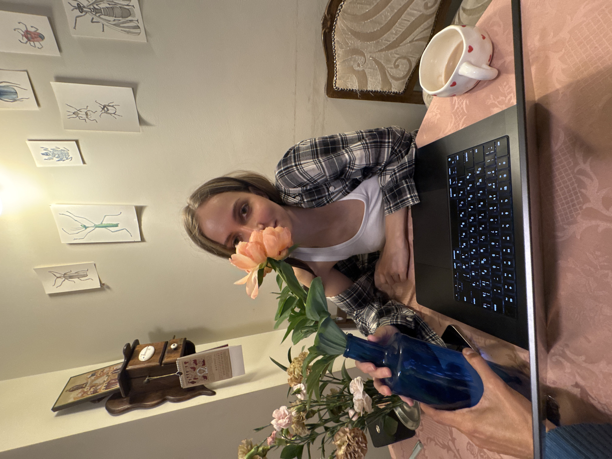 An indoor, portrait-oriented photograph captures a person in a casual setting, possibly at home in Moscow, Russia. The central figure is a young woman with light brown hair, dressed in a white tank top beneath a plaid shirt, looking directly at the viewer. She holds a light pink/peach peony flower close to her face with her left hand, appearing to appreciate its scent. Her right hand grasps a vibrant blue glass bottle containing a small arrangement of light pink and dried-looking flowers with green foliage.
She is seated at a table draped with a light pink patterned tablecloth. An open laptop, featuring a backlit keyboard with Cyrillic characters, is positioned on the table to her right. A white mug adorned with small red hearts is also visible on the table near the laptop.
The background wall is light-colored and decorated with several small, unframed drawings of various insects, including a praying mantis, beetles, and a fly. Below these, on a shelf, there is a decorative wooden box or music box with a small calendar or notepad attached. Part of an ornate chair with light-colored upholstery is visible in the upper right. The lighting is consistent with an indoor environment, likely under artificial illumination. The Cyrillic keyboard reinforces the given location context of Moscow, Russia. The scene conveys a sense of personal calm and appreciation for the flowers.