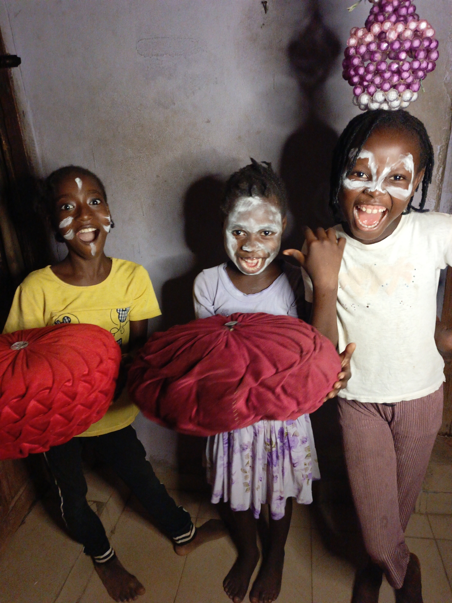 Three young girls with white face paint on their faces are captured indoors, displaying expressions of joy and excitement. The girl on the left wears a yellow t-shirt and black pants, holding a red, round cushion. Her mouth is open in a wide smile, and her eyes are wide with excitement. The middle girl, in a light purple floral dress, holds a dark red cushion and also smiles broadly with an open mouth. The girl on the right, with braided hair, wears a white t-shirt and striped brown pants, sharing a similar wide, joyful expression with an open mouth. All three are looking directly towards the camera.
The setting appears to be a dimly lit room with a textured, light-colored wall behind them and a tiled floor. A distinct large shadow, resembling a person, is visible on the wall behind the middle girl, and other shadows are cast by the subjects, suggesting an indoor setting with artificial or limited natural light. In the upper right corner, a decorative cluster of small, spherical purple and white objects, possibly plastic beads or small fruits, hangs, likely from the ceiling or an overhead fixture. The overall atmosphere is playful and energetic. There is no visible text in the image.