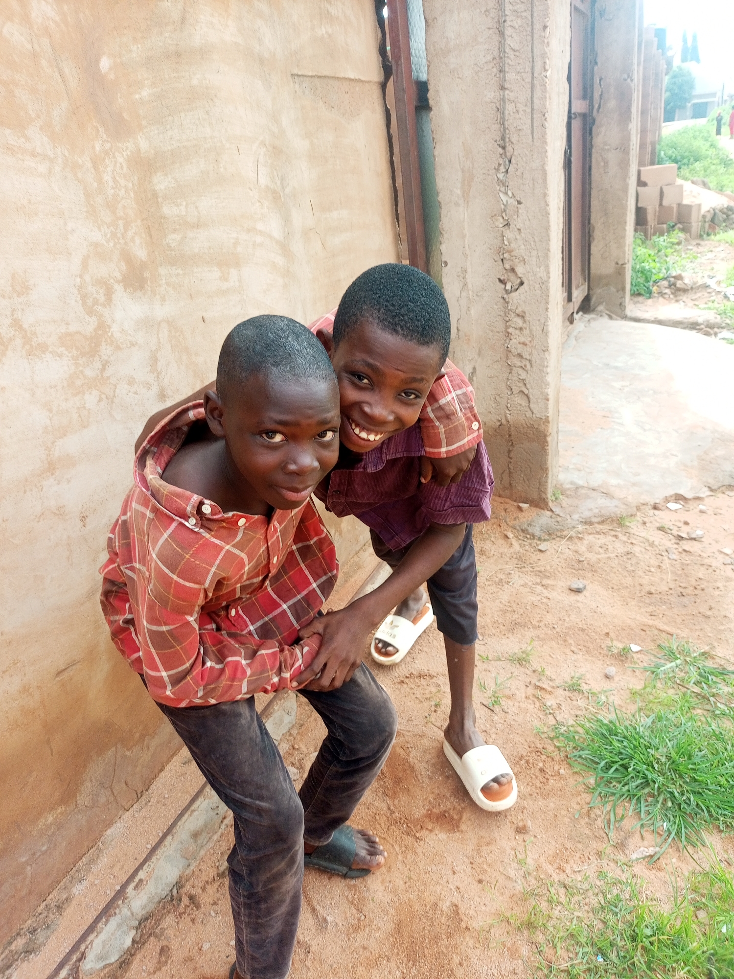 Two young boys are posed for a photo in front of a textured, tan-colored wall. The boy on the left, facing the camera, wears a red and white plaid shirt and dark pants. The boy on the right has his arm around the other's shoulders and is wearing a maroon shirt and dark pants. Both boys are smiling. The ground is dirt with patches of green grass. In the background, there's a portion of a building with a metal door and some stacked bricks. This scene is located in Tempure, Nigeria.