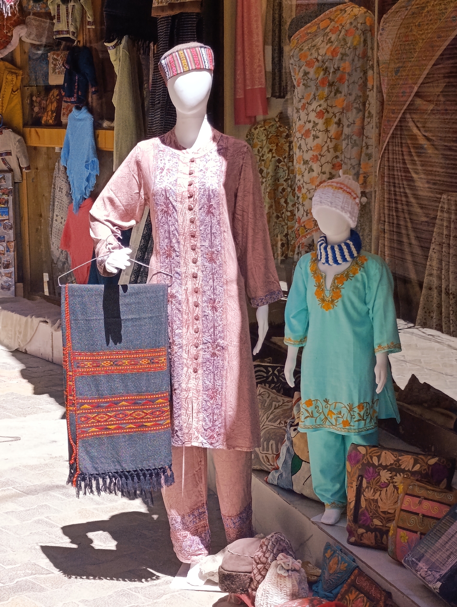 This image depicts a street-side market stall in Leh, India, likely a shop selling traditional Ladakhi clothing and accessories. The scene features two mannequins dressed in ethnic wear, attracting potential customers.
The mannequin on the left, a taller adult-sized form, is adorned in a light pink, embroidered dress with a long, button-down front and a matching patterned fabric on the lower legs, suggesting a complete outfit. It wears a traditional woven headband with colorful geometric patterns. A hanger holding a dark grey shawl with vibrant red and yellow woven stripes and tasseled edges is suspended in front of the mannequin, adding a decorative element.
To the right, a smaller, child-sized mannequin is dressed in a bright turquoise kurta with intricate yellow floral embroidery around the neckline and sleeves, paired with matching turquoise trousers. It wears a white knitted cap and a striped blue and white scarf around its neck.
The background is filled with various textiles, including patterned fabrics draped and hung, suggestive of the merchandise offered. The setting appears to be outdoors, with sunlight casting shadows on the paved ground in the foreground. The overall impression is of a vibrant and colorful marketplace typical of tourist destinations in Ladakh.