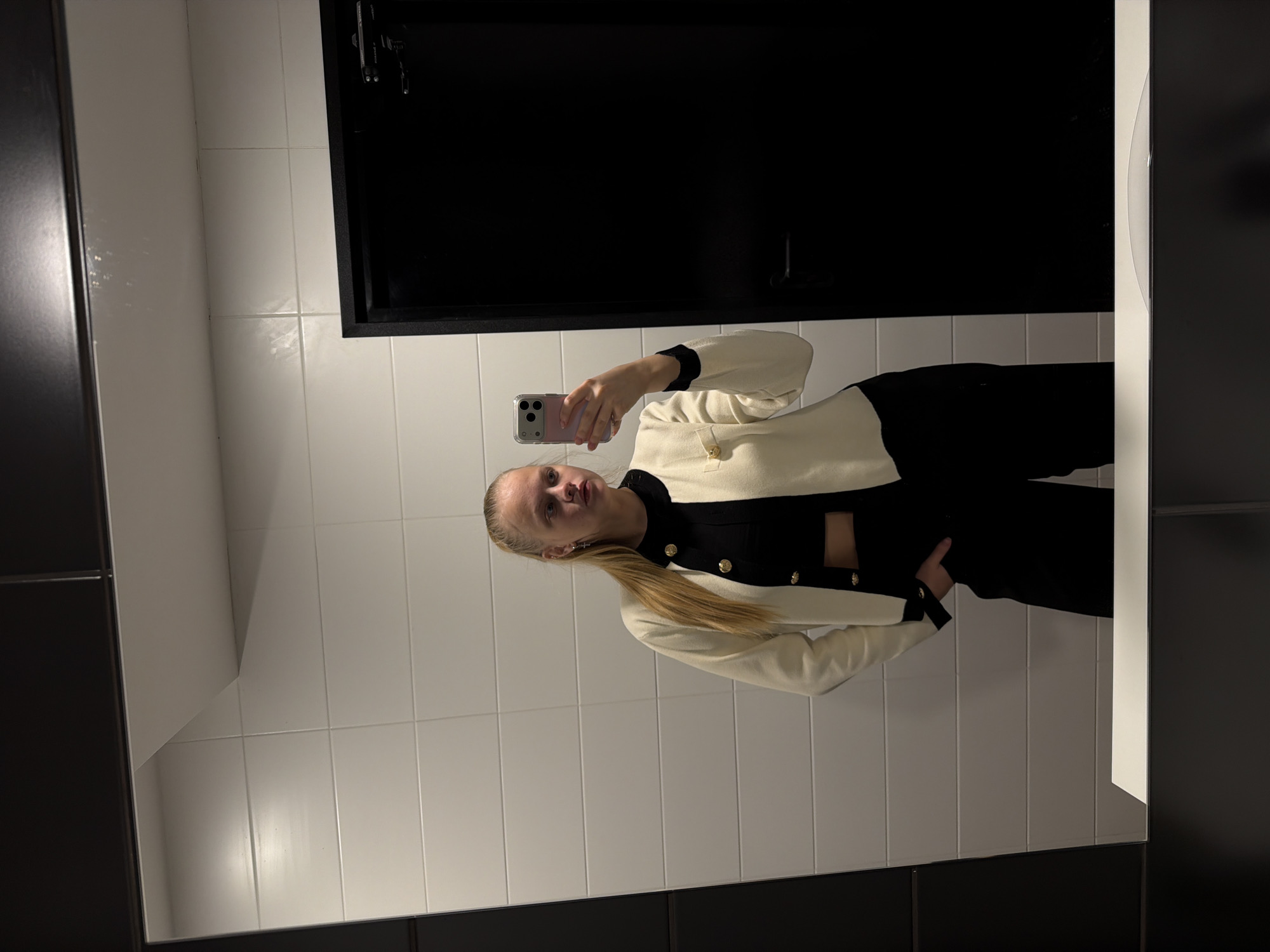 A woman is taking a selfie in front of a mirror in a bathroom. The bathroom has white tiled walls and a black door or cabinet. The woman is wearing a black and white outfit. Her hair is blonde and pulled back in a ponytail. There is no visible evidence indicating the city of Izhevsk, Russia.