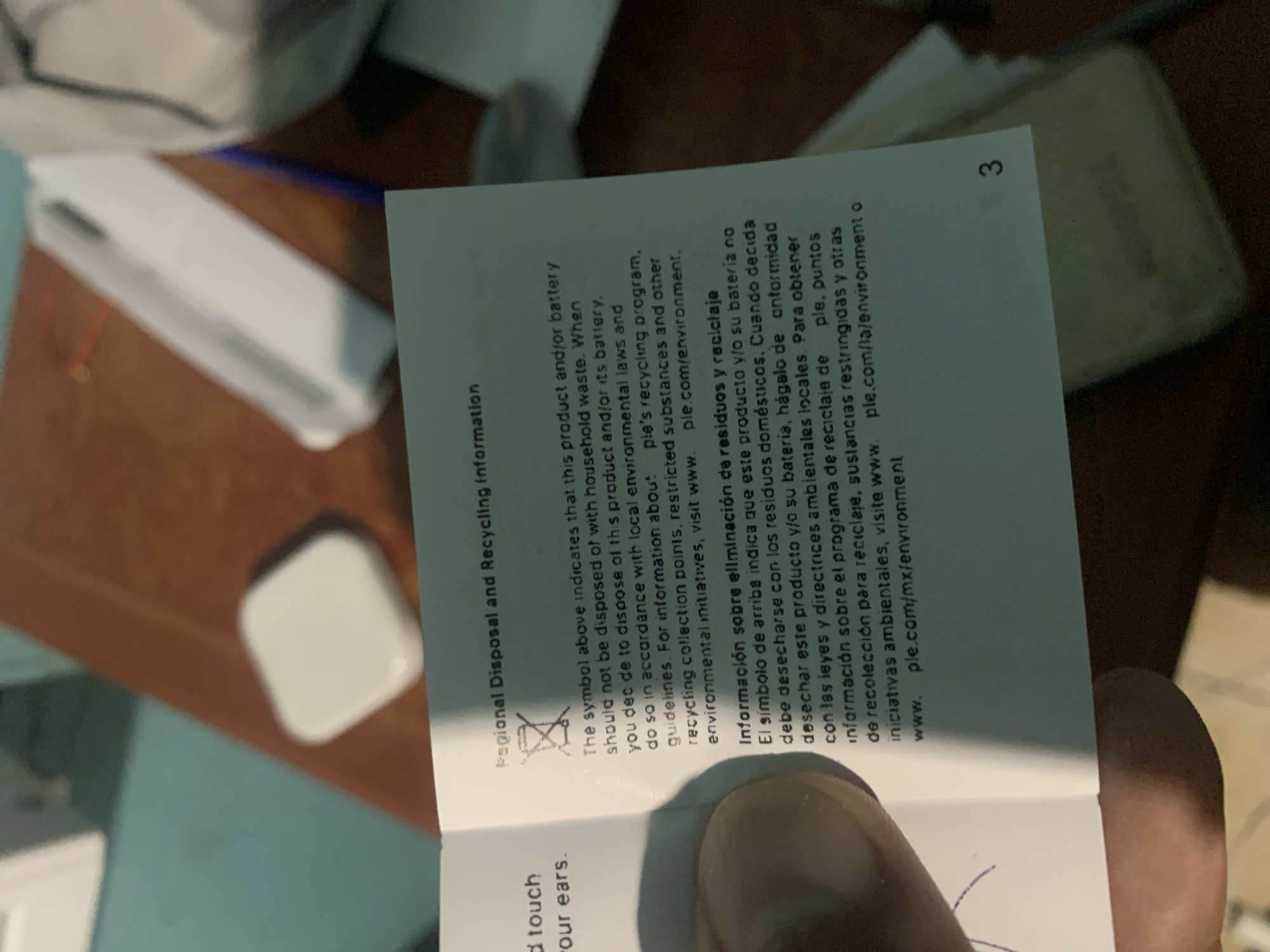 The image captures a close-up of a person's hand holding open a small white information booklet. The booklet is titled "Regional Disposal and Recycling Information" and features text in both English and Spanish.
The visible English text states that the product and/or battery "should not be disposed of with household waste" and advises disposal "in accordance with local environmental laws and guidelines." It directs users to "www. ple.com/environment" for details on the manufacturer's recycling program and environmental initiatives. The Spanish text provides similar instructions regarding waste disposal and recycling, mentioning "www.ple.com/la/environment o www.ple.com/mx/environment". A crossed-out wheeled bin symbol, indicating that the item should not be discarded with general waste, is clearly visible. The page number '3' is present in the top right corner of the page. A small portion of text, "touch our ears.", is visible at the bottom left, partially obscured by the thumb.
In the background, a blurry white electronic device, possibly a charging case or a similar small accessory, rests on a light brown wooden surface. A blue surface is also visible underneath the booklet, suggesting an indoor setting, likely a table or desk. The activity shows an individual reviewing product information regarding environmental disposal.