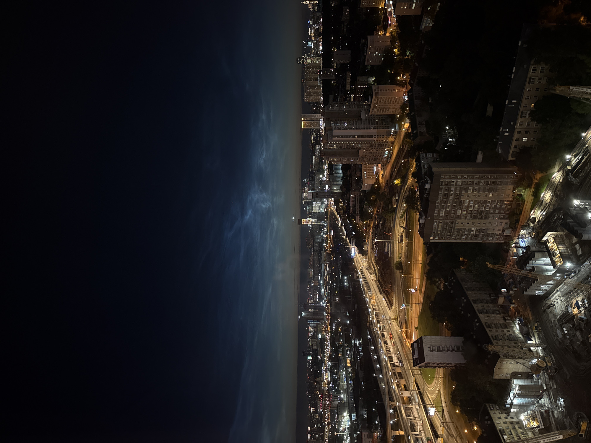 The image shows a nighttime aerial view of the city of Khimki, Russia. The sky is dark with visible noctilucent clouds, appearing as wispy, luminescent streaks. Below the clouds, the city is illuminated by a multitude of lights from buildings and streetlights. A complex network of elevated roads and highways is prominent in the mid-ground, with cars visible on them. Several high-rise apartment buildings and office buildings are scattered throughout the cityscape. In the foreground, a construction site is visible with cranes and unfinished structures, indicating ongoing development. The overall scene depicts a bustling urban environment at night, with a striking atmospheric phenomenon in the sky.