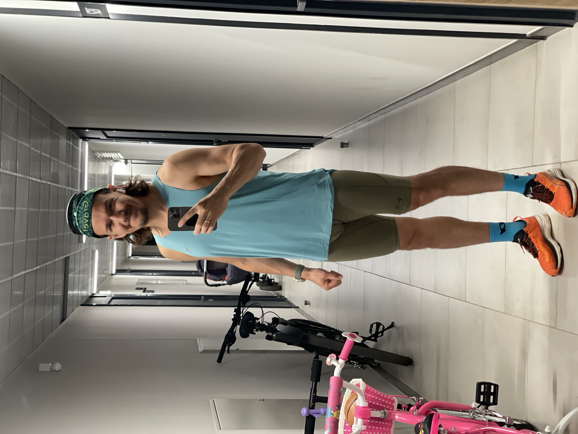 A man with long brown hair and a beard stands in a brightly lit hallway, taking a mirror selfie. He is wearing a teal tank top, olive green compression shorts, blue athletic socks, and orange running shoes. He has a green bandana on his head. He is holding a black smartphone in his right hand and wearing a watch on his left wrist. To his left, there is a black bicycle and a pink child's bicycle. In the background, a stroller is visible. The hallway appears to be in a residential building, with white walls and a tiled floor. The ceiling has fluorescent lighting.
