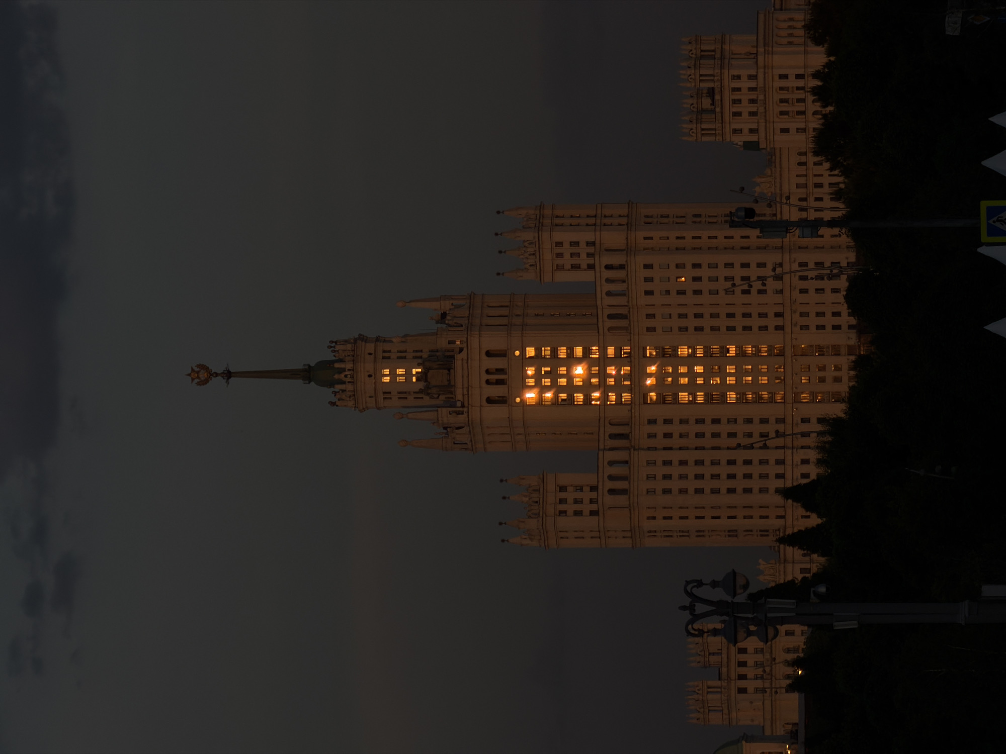 The image depicts one of the Seven Sisters skyscrapers in Moscow, Russia, illuminated by the setting sun. The building is a Stalinist-era architectural landmark with a central tower and ornate detailing. The facade is marked by rows of windows, some of which reflect a golden light, contrasting with the dark sky and the surrounding shadows from trees and street lighting. The scene appears to be at dusk or early evening.