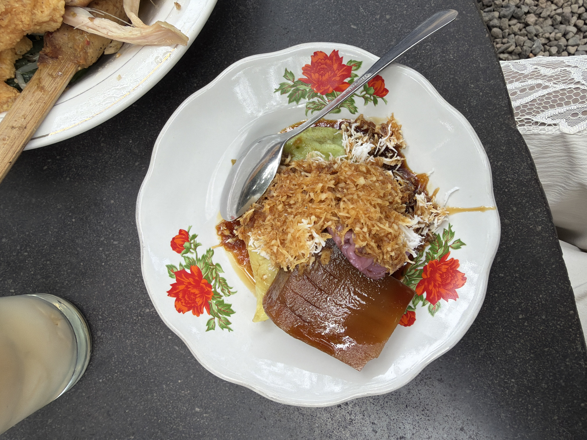 A close-up overhead shot captures a plate of Indonesian food, likely a dessert, served on a white ceramic plate with a floral pattern. The plate is scalloped around the edges and decorated with vibrant red flowers and green leaves. The food itself appears to be a layered confection, with a dark, translucent jelly-like substance at the bottom. Above this, there's a light green creamy layer, followed by a mound of shredded, toasted coconut mixed with some darker, syrupy sauce. Underneath the coconut and sauce, hints of purple are visible, possibly from sweet potato or taro. A silver spoon rests on the plate, partially submerged in the food, suggesting it's ready to be eaten.
In the background, to the left, a portion of another plate is visible, containing what looks like fried food items on skewers. To the right of the main plate, a white lacy fabric is draped, hinting at a tablecloth or clothing. A glass filled with a pale, creamy liquid is also visible in the bottom left corner of the frame. The setting appears to be outdoors, with a dark, textured surface beneath the plates, possibly a stone table, and small grey pebbles visible in the upper right background. The lighting suggests it's daytime, with no indication of specific weather conditions. The overall mood is casual, perhaps a meal enjoyed outdoors in a relaxed setting. The location is identified as Kuta, Indonesia. There is no visible text in the image.