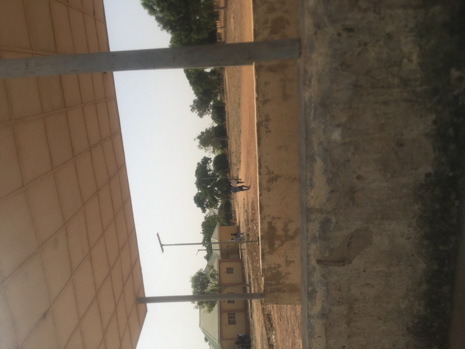 The image displays an outdoor scene in Pindiga, Nigeria, captured from beneath a light-colored, gridded overhead canopy supported by two grey metal poles. A weathered concrete barrier, exhibiting discoloration, cracks, and dark staining, spans the lower foreground. Beyond this barrier, a dry expanse of reddish-brown dirt or sandy ground extends into the midground. Approximately five to seven individuals are observed walking in small groups across this open ground, moving generally from left to right. To the left, two single-story buildings with light yellow walls and green hip roofs are visible. Two tall, grey poles topped with solar streetlights are positioned in the midground. The background consists of scattered green trees, and the sky is a hazy, pale blue.