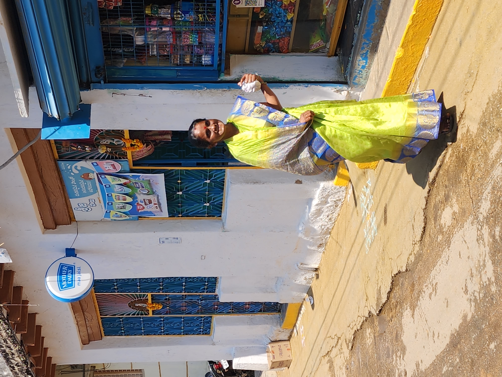 An adult female stands on a concrete path in front of a white-painted building in Pedda Kammavaripalli, India, under direct sunlight. She is positioned slightly to the right of the frame's center, facing the viewer with a smile. She wears a lime green sari with blue and gold borders and dark sandals, and has a red bindi on her forehead. Her right hand holds a small white, crumpled object.
The building behind her features a textured white facade with a yellow painted base. To the left, a blue-framed doorway with an intricate metal grille is adorned with an orange flower garland. Above this, a circular blue and white "MOTHER DAIRY maa jaisi" sign is mounted. Further left, a partial view of a parked dark-colored motorcycle is visible next to a brown cardboard box labeled "Rocket." To the right of the woman, a storefront is visible with a partially raised blue roll-down shutter, revealing shelves stocked with numerous packaged items. A QR code sticker is affixed inside the storefront. A second blue-framed window/doorway with a grille is present to the left of the storefront. A poster with text in a local language (likely Telugu) is affixed to the wall between the two doorways. The path shows cracks and discoloration, with white chalk-like patterns on the ground near the building. Shadows are cast by the woman and the building, indicating bright overhead sunlight.