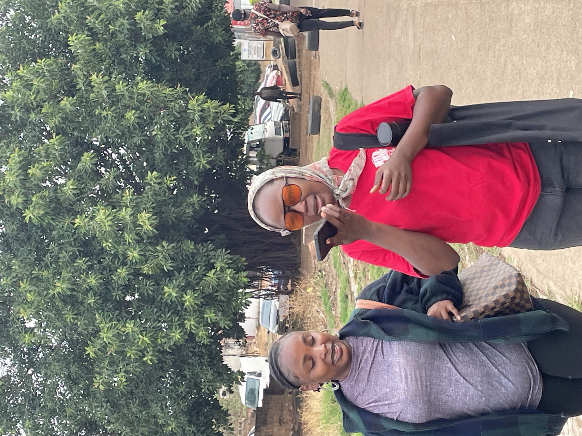 Two women are standing outside near a large green tree in Jos, Nigeria. One woman wears a red shirt, orange sunglasses, and a patterned headscarf, and she holds a smartphone. The second woman, clad in a grey shirt and a green and blue plaid jacket, has a handbag resting on her lap. Cars are parked nearby, and people can be seen in the background on a concrete walkway.