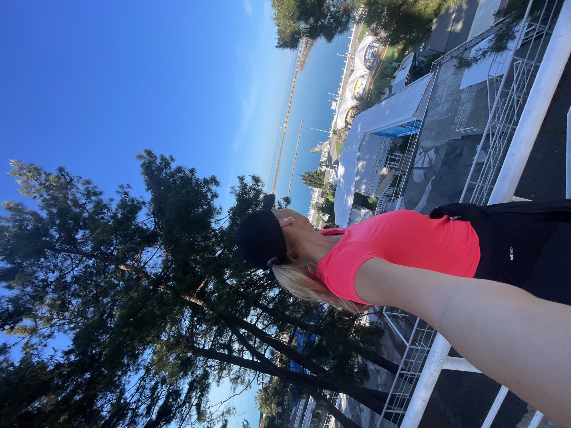 This bright, clear daytime image is a selfie taken from an elevated perspective, likely a balcony or viewing platform, overlooking a coastal scene in Sochi, Russia.
The primary subject is a woman, positioned on the right side of the frame, captured from the chest up as she holds the camera. She has blonde hair pulled into a ponytail and wears a black baseball cap, a vibrant neon pink short-sleeved top, and black athletic bottoms that bear the partially visible text "MOVING". She is looking upwards and outwards, seemingly appreciating the panoramic view.
In the mid-ground, several tall, dark green coniferous trees, possibly pines, rise from below, framing the left side of the scene and partially obscuring some of the structures directly beneath the viewpoint.
The background reveals a vast, serene expanse of deep blue sea under a clear, cloudless sky. Stretching into the distance across the water are two prominent, long breakwaters or piers, extending far out from the shore. Along the coastline, several white and light-colored modern buildings with distinctive arched roofs and facades are visible. These structures suggest a waterfront complex, possibly a marina, resort, or entertainment area.
The strong, direct sunlight indicates a sunny day, likely morning or early afternoon, with excellent visibility. The overall atmosphere conveys leisure and enjoyment of a scenic, clear day at a coastal location.