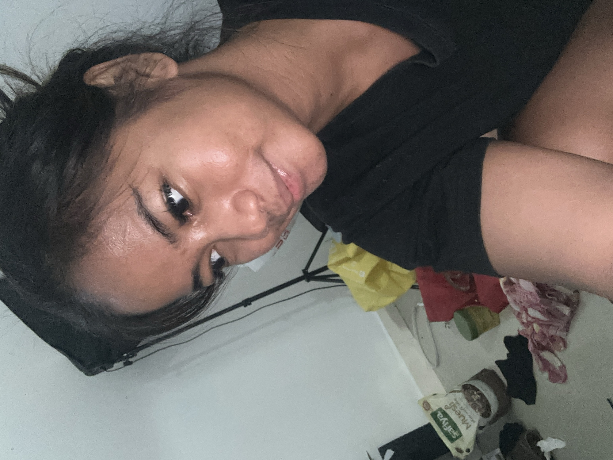 A young woman with dark hair, pulled back, is shown in a close-up selfie from the chest up, against a plain wall. She is wearing a black t-shirt and appears to be sweating. Her face is illuminated, highlighting her skin and features. In the background, a light stand is visible, along with some scattered items on the floor, including a yellow bag, a red cloth, a green container, a bag of muesli, and other fabric items. The overall setting appears to be an indoor domestic environment, possibly a room in Sukawati, Indonesia.
