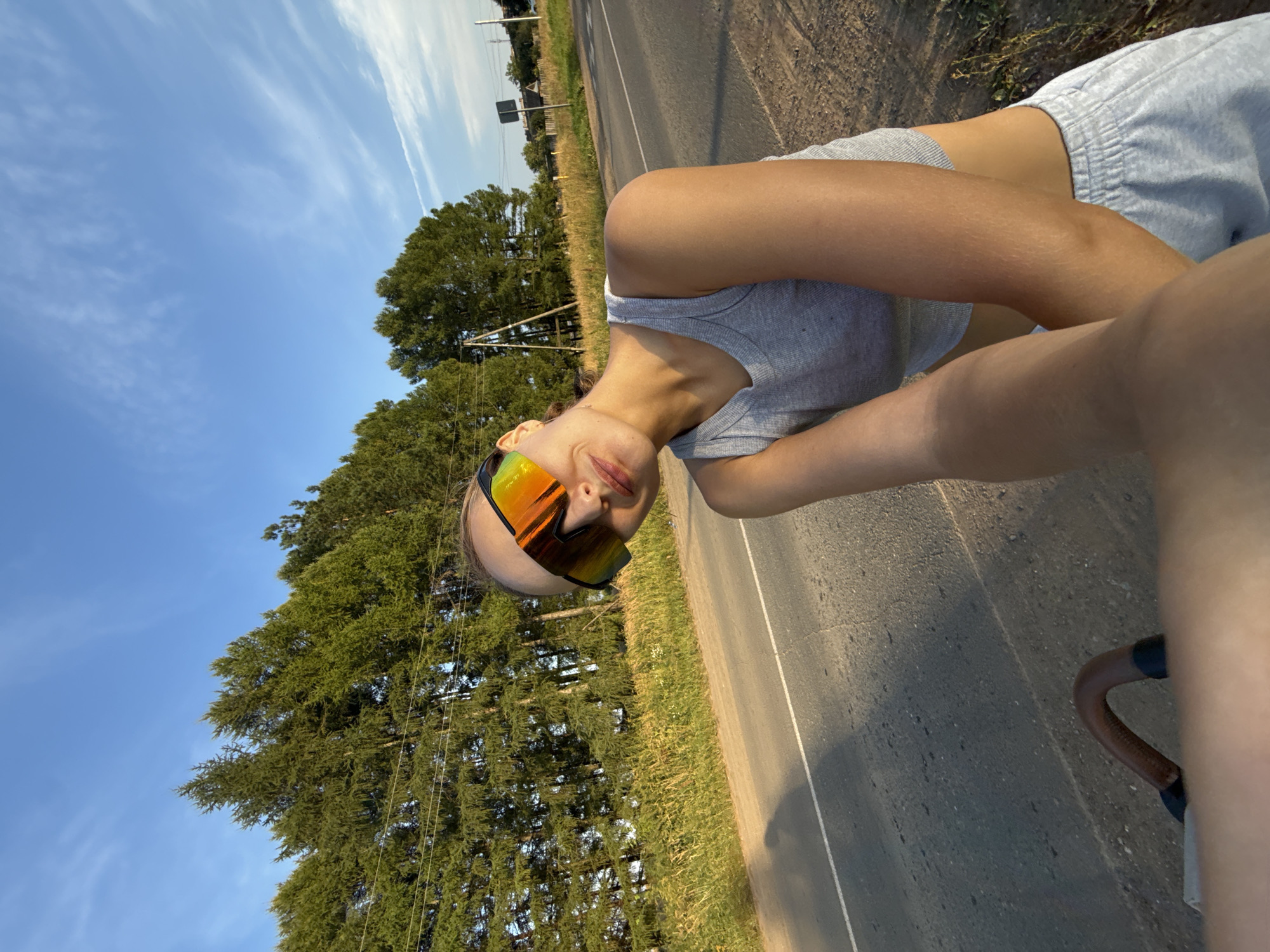 A woman wearing sunglasses and a grey tank top is captured in a selfie outdoors. She is positioned in the foreground, leaning slightly towards the camera, with a faint smile. Behind her, a paved road stretches into the distance, bordered by a grassy embankment and a dense line of evergreen trees. The sky above is a clear blue with a few wispy clouds. The scene appears to be set in a rural or suburban area, possibly on the outskirts of Подшивалово, Russia, given the natural landscape and absence of prominent urban structures. The lighting suggests it is late afternoon or early evening, with the sun casting a warm glow on the environment.