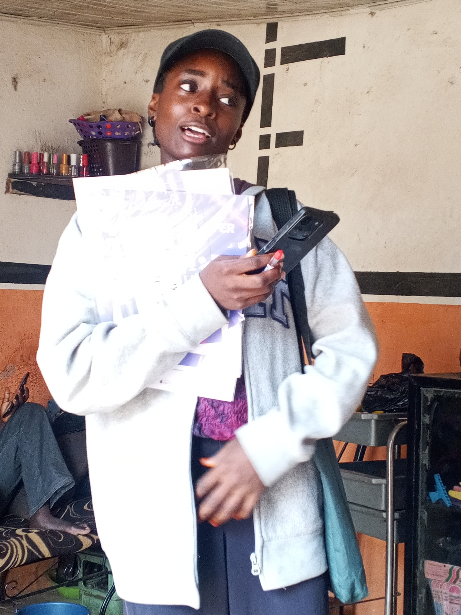 A young woman in Gwong, Nigeria, stands indoors. She wears a gray hooded sweatshirt, a dark cap, and carries papers and a smartphone. A person's legs and bare foot rest on a patterned bench behind her. In the background are shelves with nail polish and various containers. The walls are painted in beige and orange, with black painted decorative lines. A utility cart sits to the right.