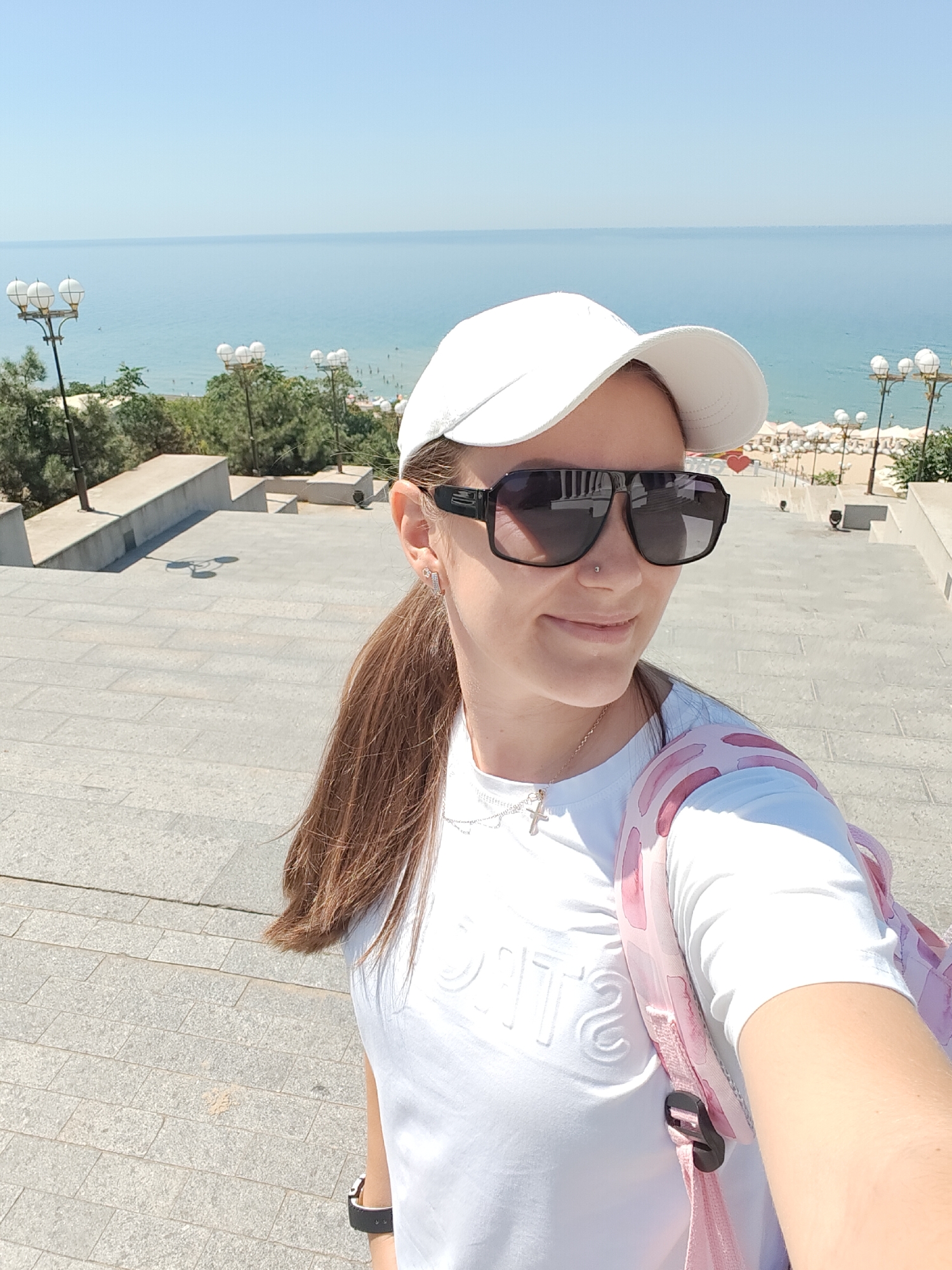 A woman wearing a white baseball cap and oversized sunglasses stands on a stone staircase, looking to her right and smiling. She has long brown hair pulled back in a ponytail and is wearing a white t-shirt with embossed lettering. A pink backpack with a watercolor-like pattern is slung over her left shoulder. In the background, the Black Sea stretches out under a clear blue sky, with a beach lined with white umbrellas visible in the distance. Streetlights and trees are scattered along the upper portions of the staircase and embankment. The scene appears to be a public area in Chornomorsk, Ukraine, likely a promenade or viewing area overlooking the sea.