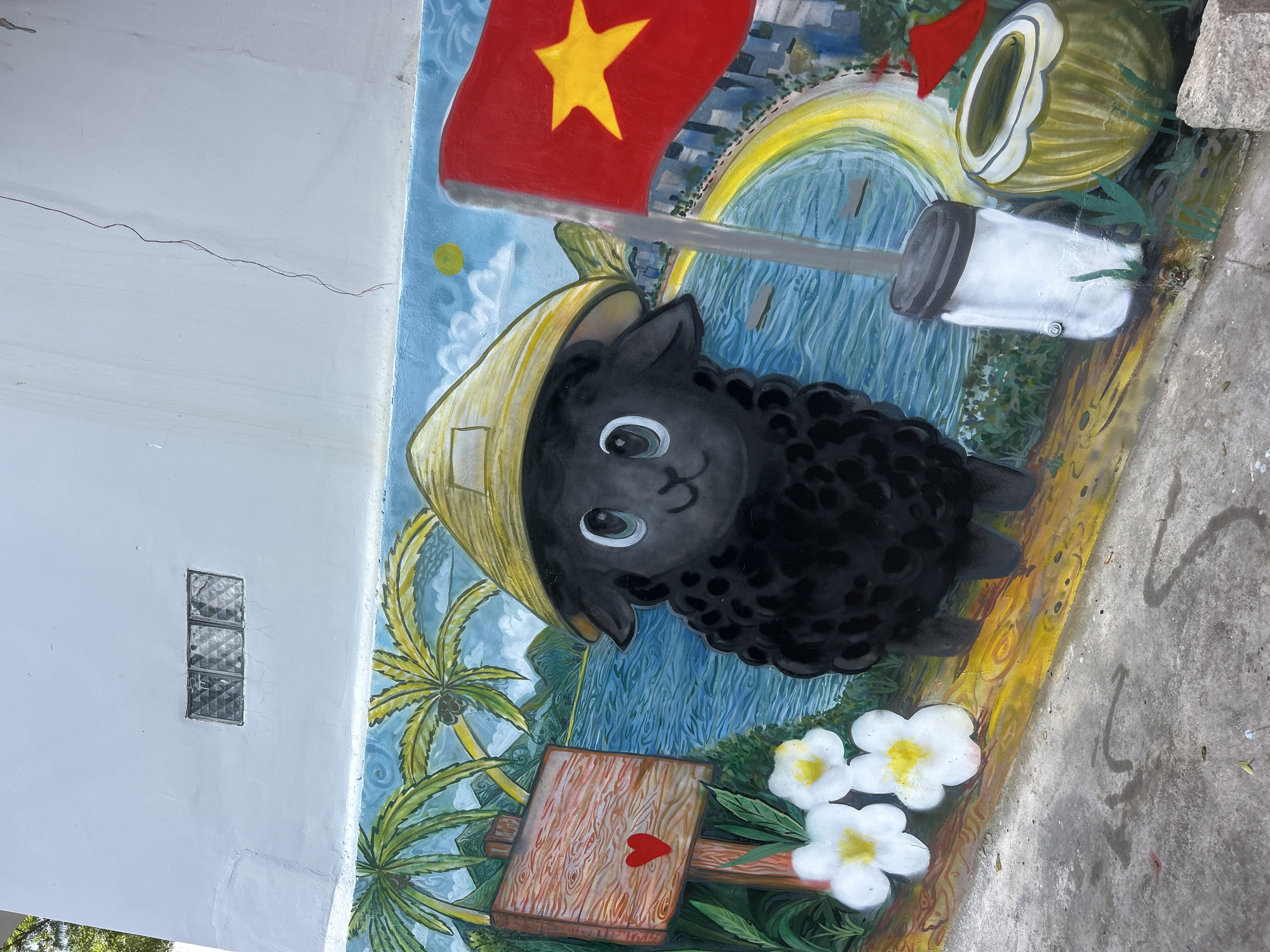 This image depicts a mural in Bac Nha Trang Ward, Vietnam. The mural is painted on a white wall bordering a concrete surface. The mural features a black sheep wearing a traditional conical hat. Behind the sheep, there is a background depicting a city skyline, a yellow arch, a body of water, and a coconut. The Vietnamese flag is prominently displayed with a yellow star. Additionally, there are palm trees, flowers, and a wooden mallet with a red heart on it.