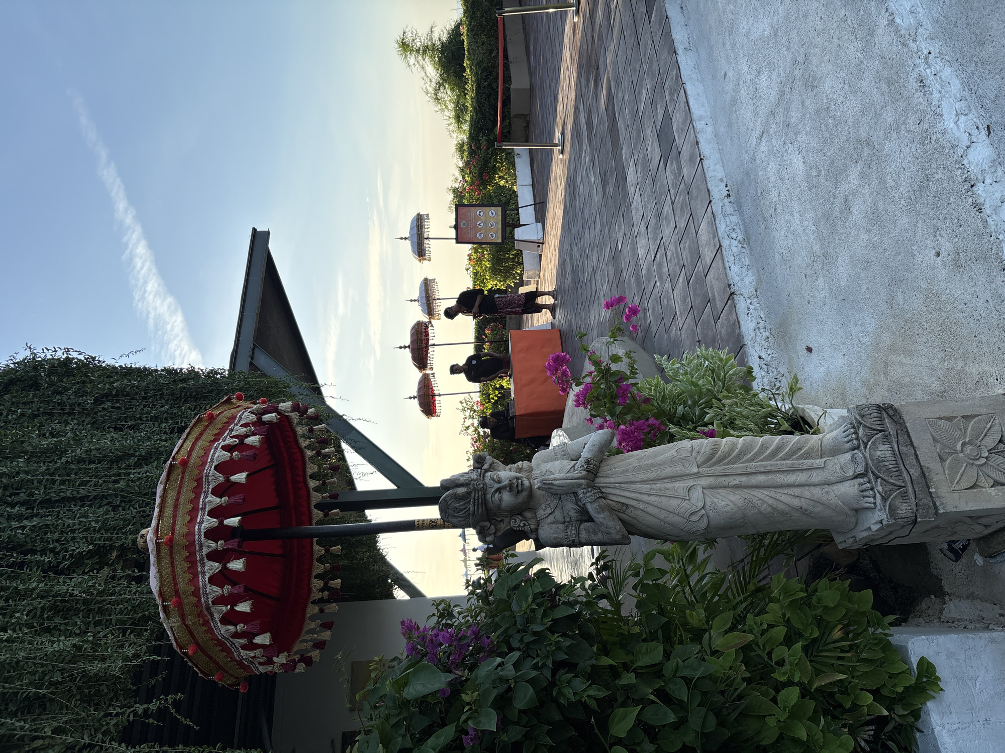 An outdoor scene at dusk shows a welcome statue in the foreground, with lush green foliage to its left and pink flowers. The statue is a carved figure with its hands pressed together in a prayer-like gesture. Behind the statue, a paved walkway leads to a reception area where two men in traditional attire are standing. An orange table is set up, and several decorative umbrellas are displayed. In the distance, the ocean is visible, and the sky is a clear blue with a few wispy clouds. The overall ambiance is serene and welcoming, suggesting a tropical resort or cultural venue.