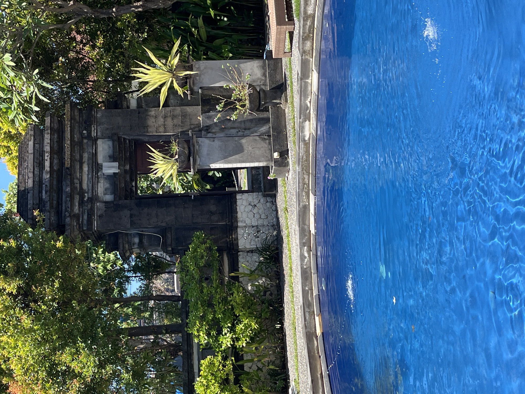 A daytime shot depicts an outdoor scene in Kuta Selatan, Indonesia. On the left, a multi-tiered stone structure, possibly a traditional Balinese temple or building facade, is partially obscured by lush greenery. To the right, a blue-tiled swimming pool reflects sunlight. The structure and pool are separated by a stone border, with plants and foliage interspersed. The water's surface shows gentle ripples, suggesting movement. The atmosphere is sunny and tropical. No people are visible.